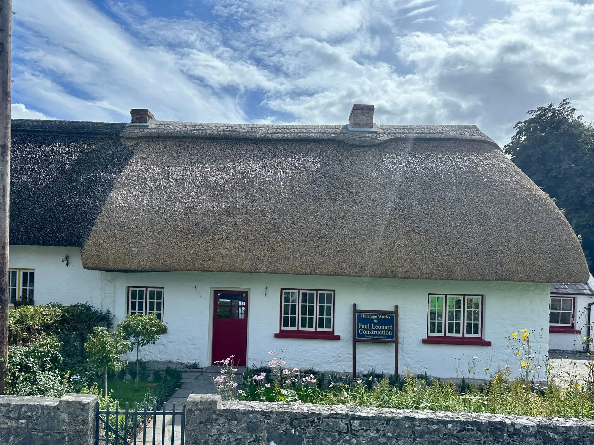 Weiss gekalktes Haus mit dickem Reetdach und roten Fensterrahmen in Adare, davor ein Blumengarten.