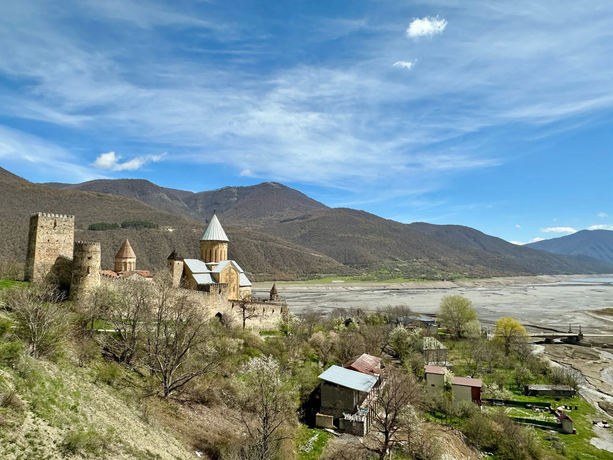 Der Festungs- und Kirchenkomplex von Ananuri mit steinernen Türmen und Kirchen vor kahlen Bergen und einer breiten Wasserfläche.