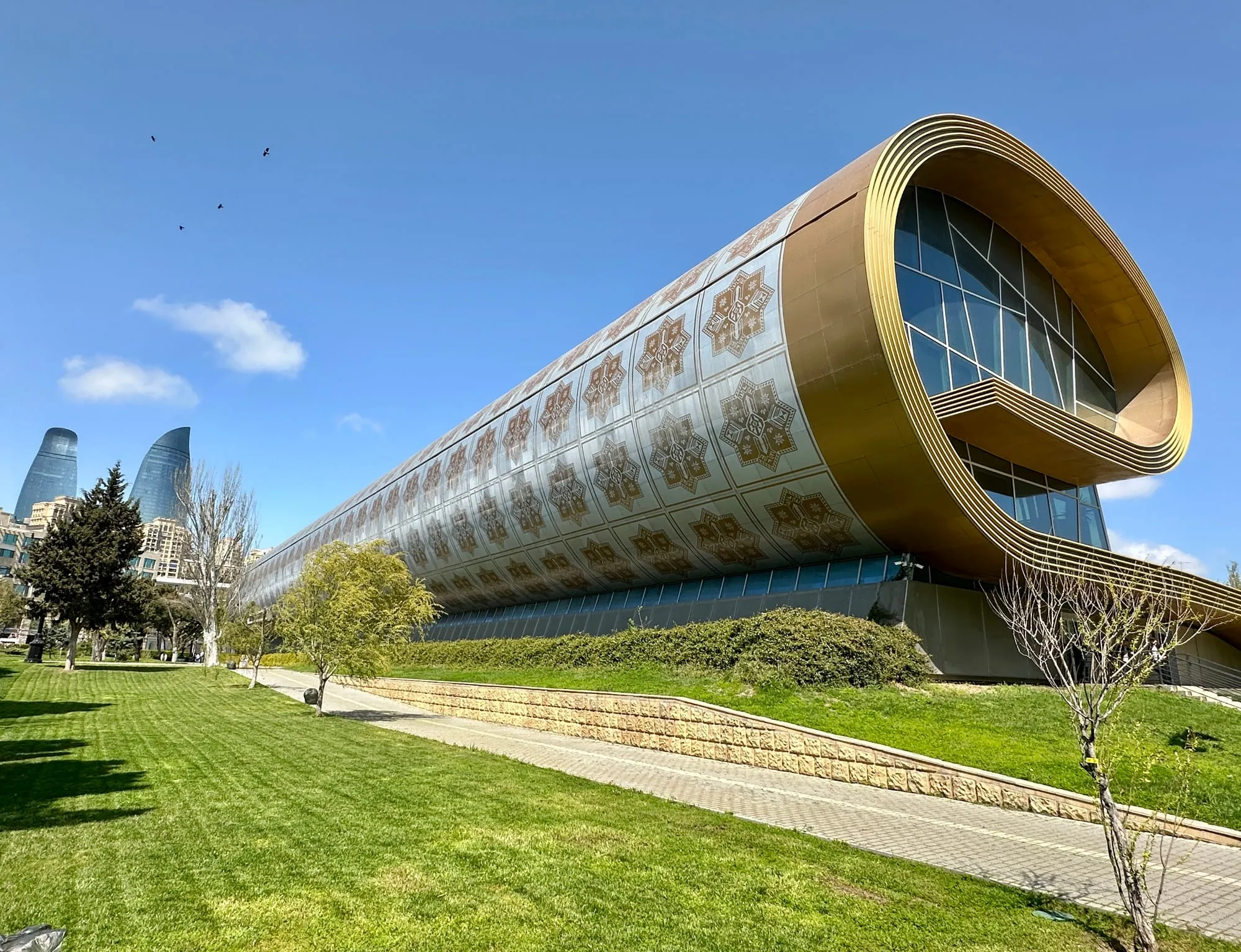 Geschwungene Fassade des Aserbaidschanischen Teppichmuseums mit geometrischen Teppichmustern, links im Hintergrund die Flame Towers über einem Park.