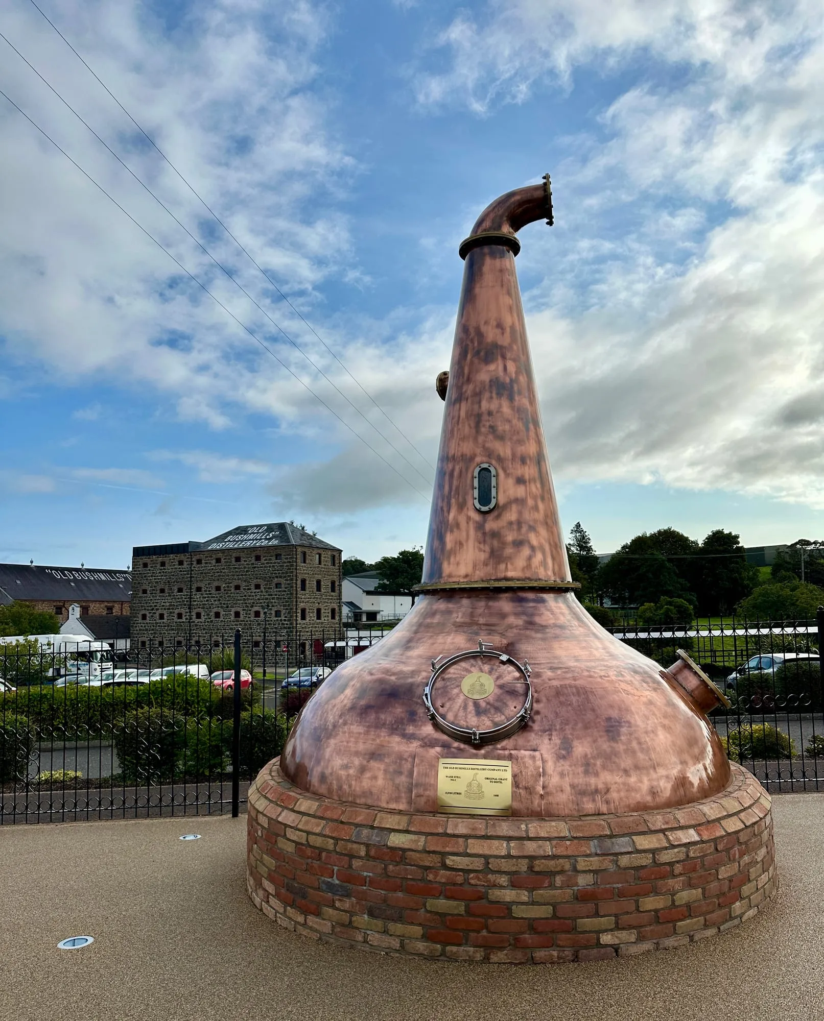 Grosser kupferner Brennkessel als Denkmal vor den Steingebäuden der Old Bushmills Distillery.