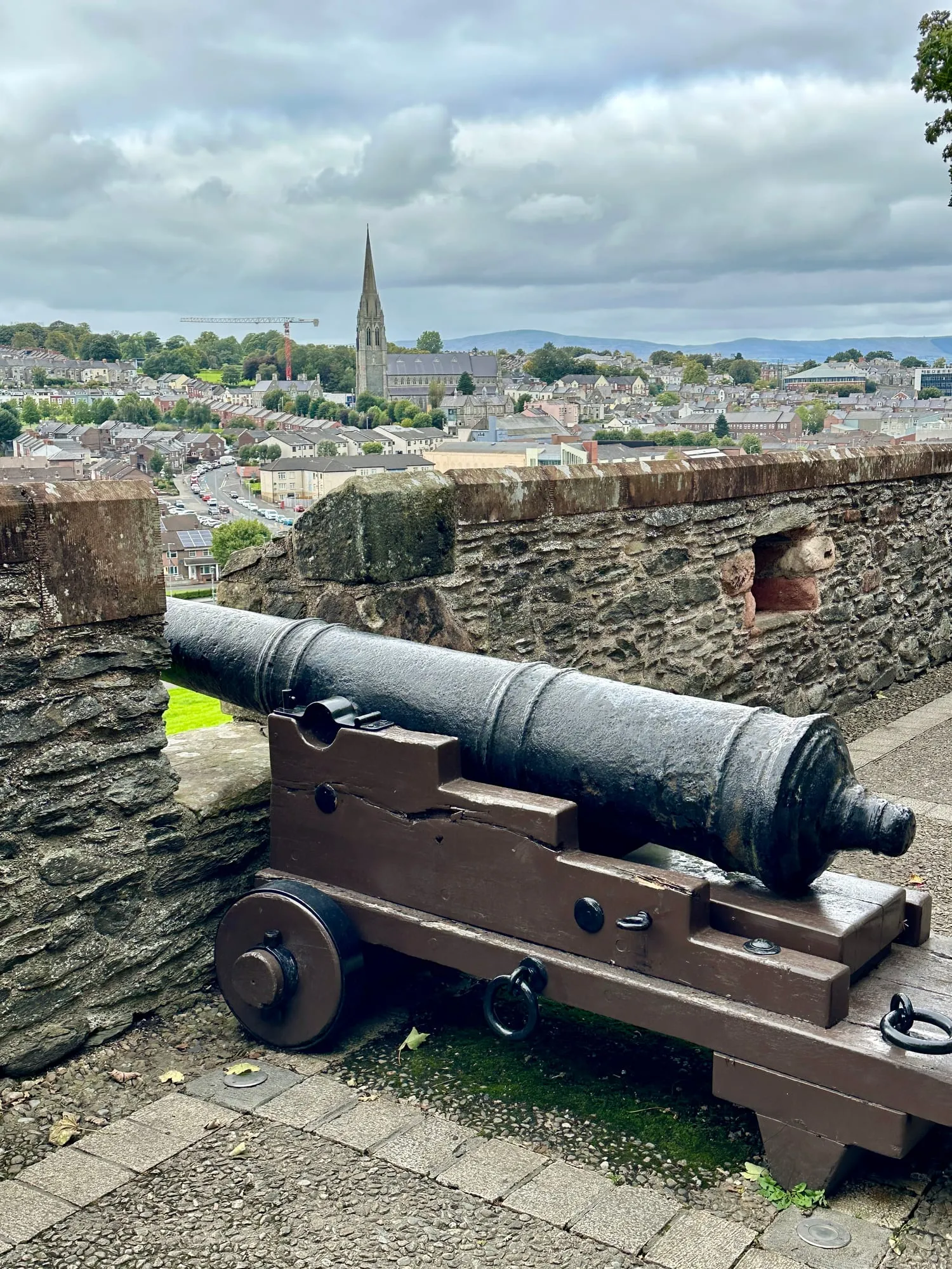 Historische Kanone auf der Stadtmauer von Derry, dahinter der Blick über die Dächer des Bogside zur Kathedrale.