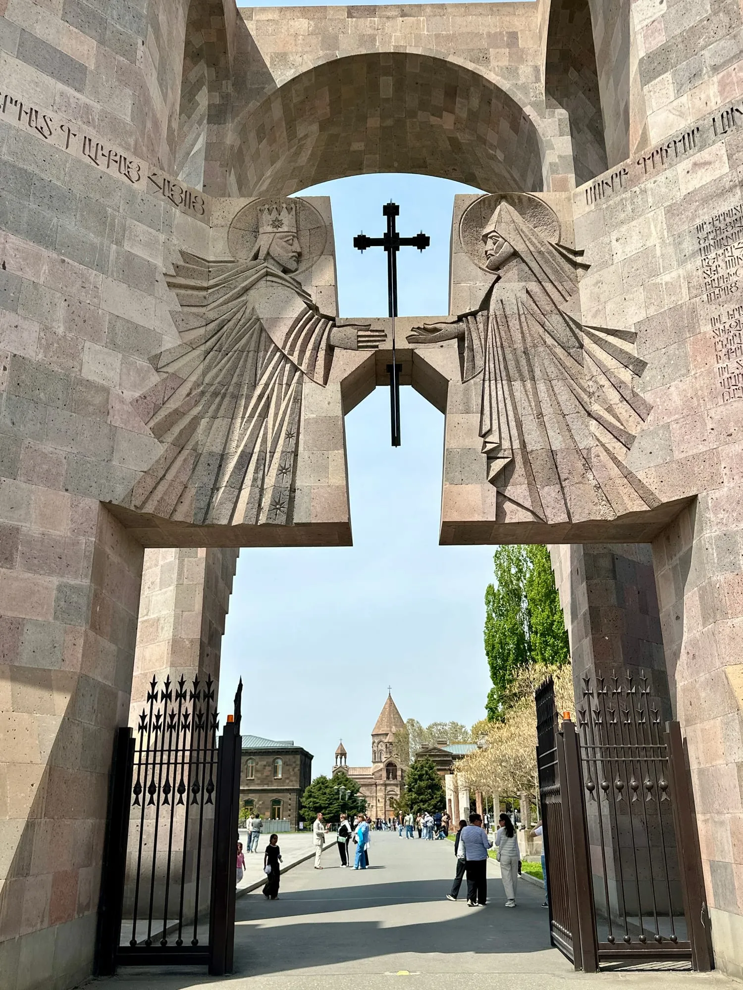 Monumentales Steintor mit Kreuz und armenischen Reliefs, dahinter eine Kirche und Besucher in Etschmiadsin.
