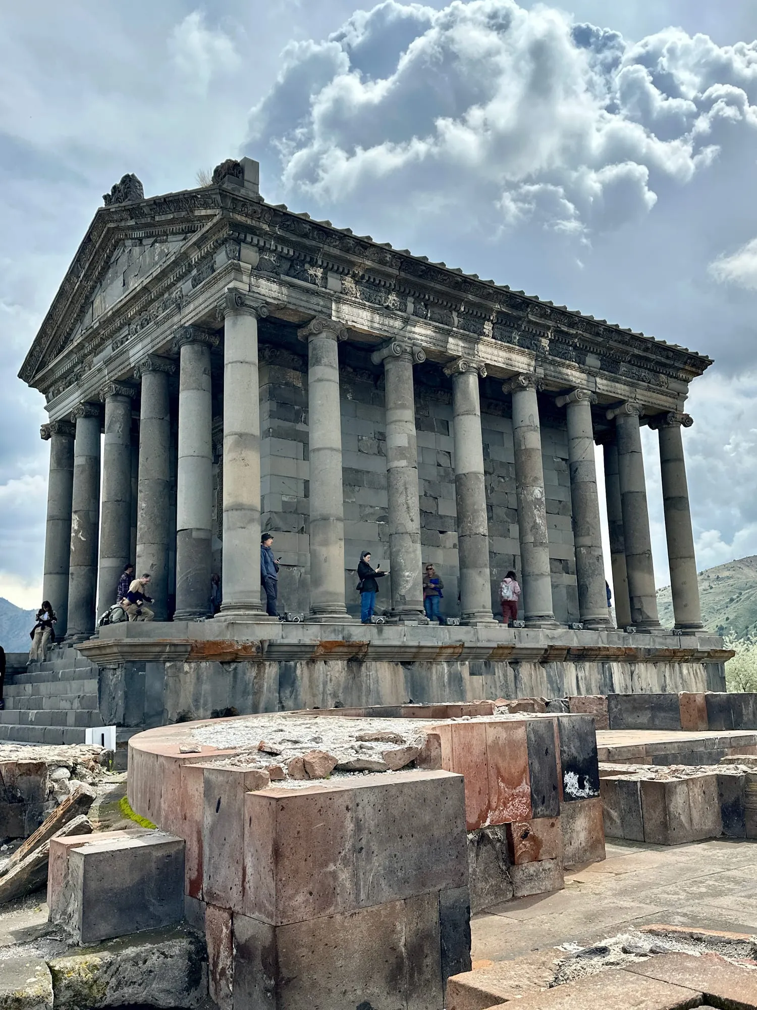 Nahaufnahme des Tempels von Garni mit hohen Säulen und reich verziertem Gebälk vor einem Wolkenhimmel, Besucher stehen in der Vorhalle.