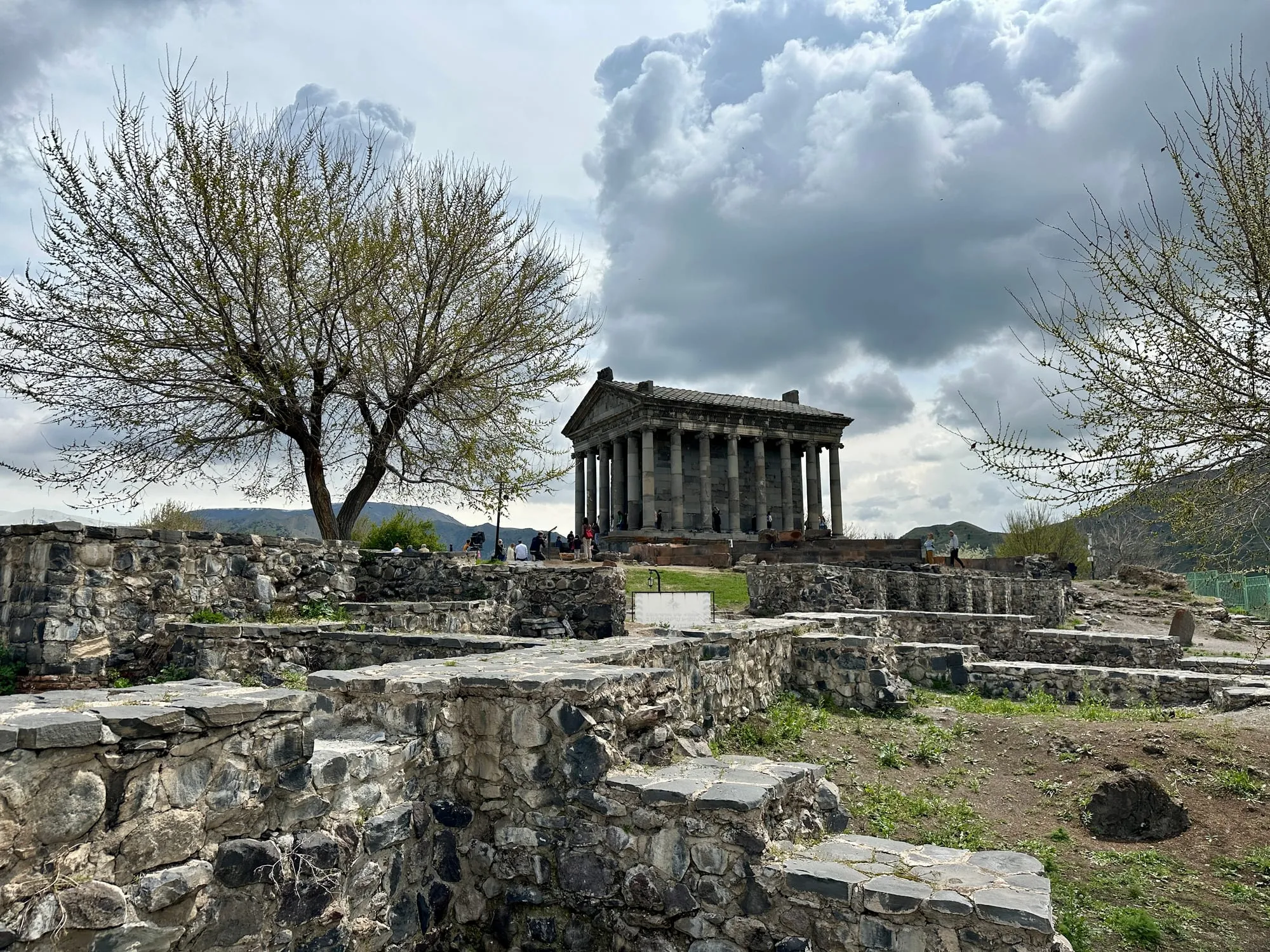 Der Garni-Tempel mit Säulen und Dreiecksgiebel steht zwischen steinernen Ruinen, gerahmt von einem grossen Baum und Hügeln unter einem wolkenreichen Himmel.