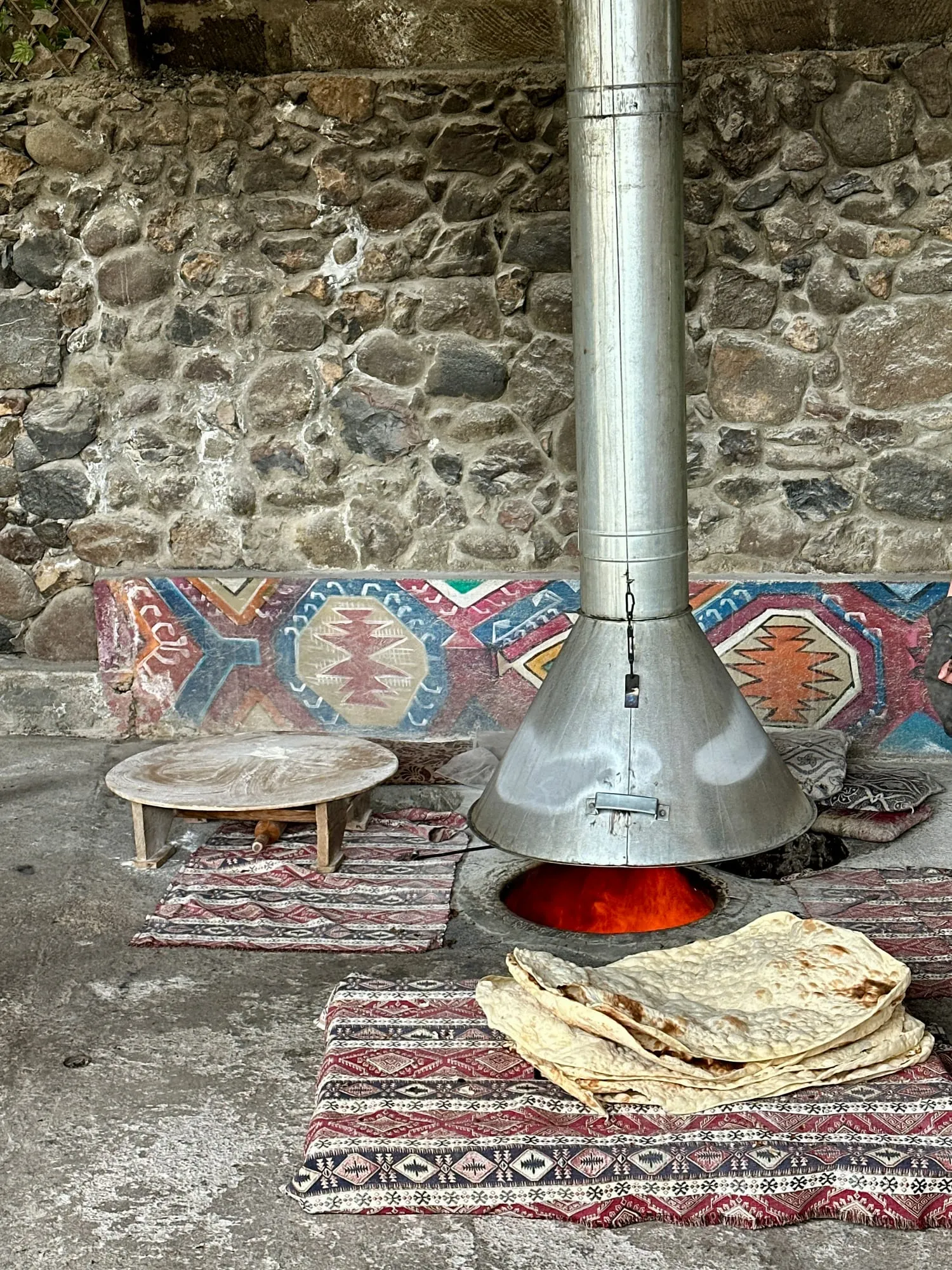 In den Boden eingelassener Backofen mit rot glühender Öffnung und Blechabzug, davor gestapelte Lavash-Fladen auf gemusterten Teppichen vor einer Natursteinmauer.