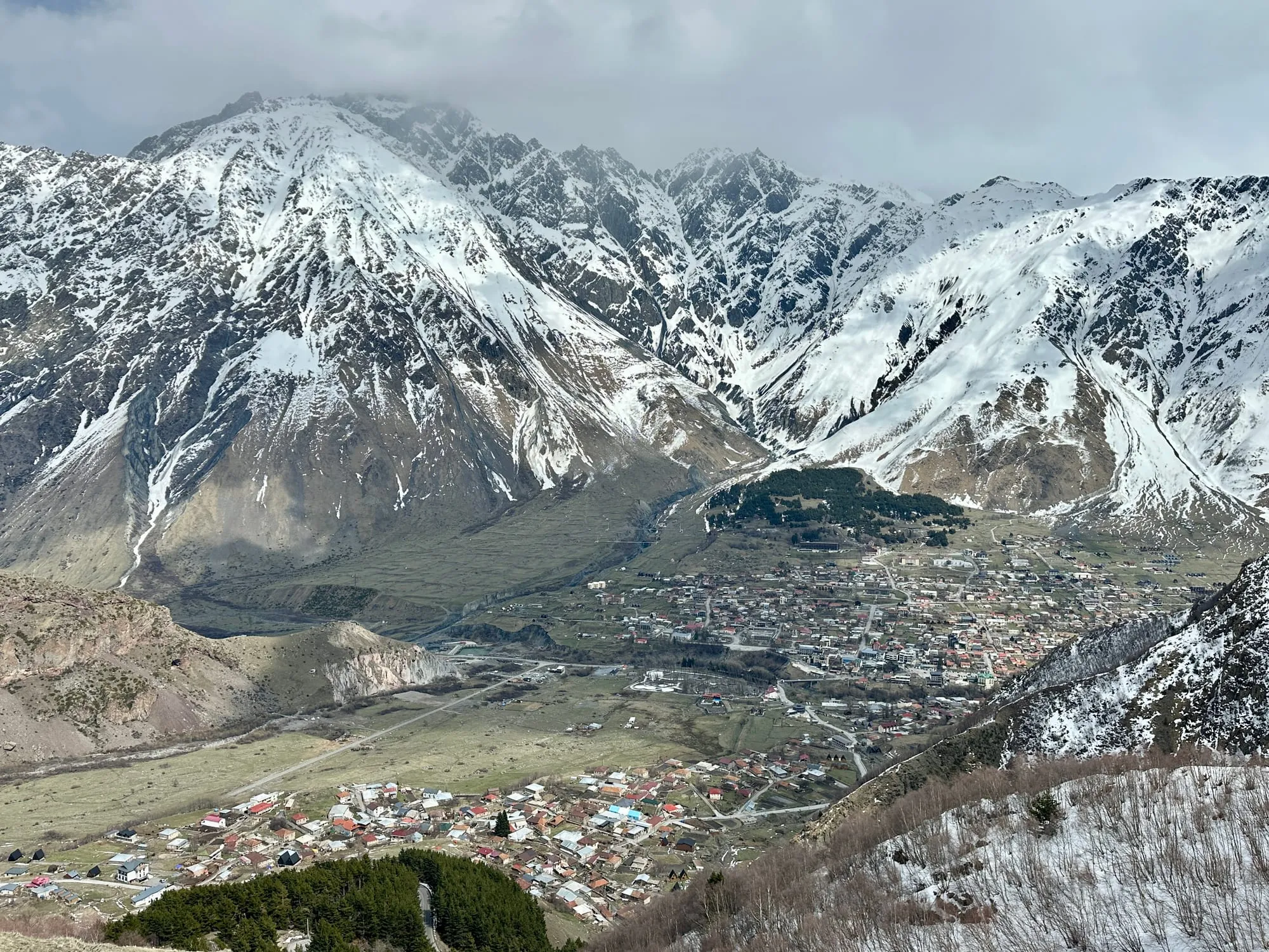 Schroffe, teils verschneite Kaukasusberge über einer Siedlung und grünen Hängen in einem Hochtal.