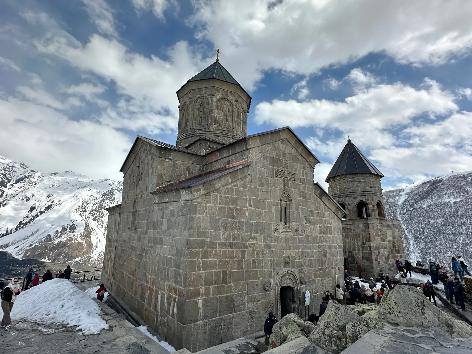 Graue Dreifaltigkeitskirche von Gergeti mit Kreuzen vor verschneiten Berghängen, Besucher stehen auf dem felsigen Vorplatz.