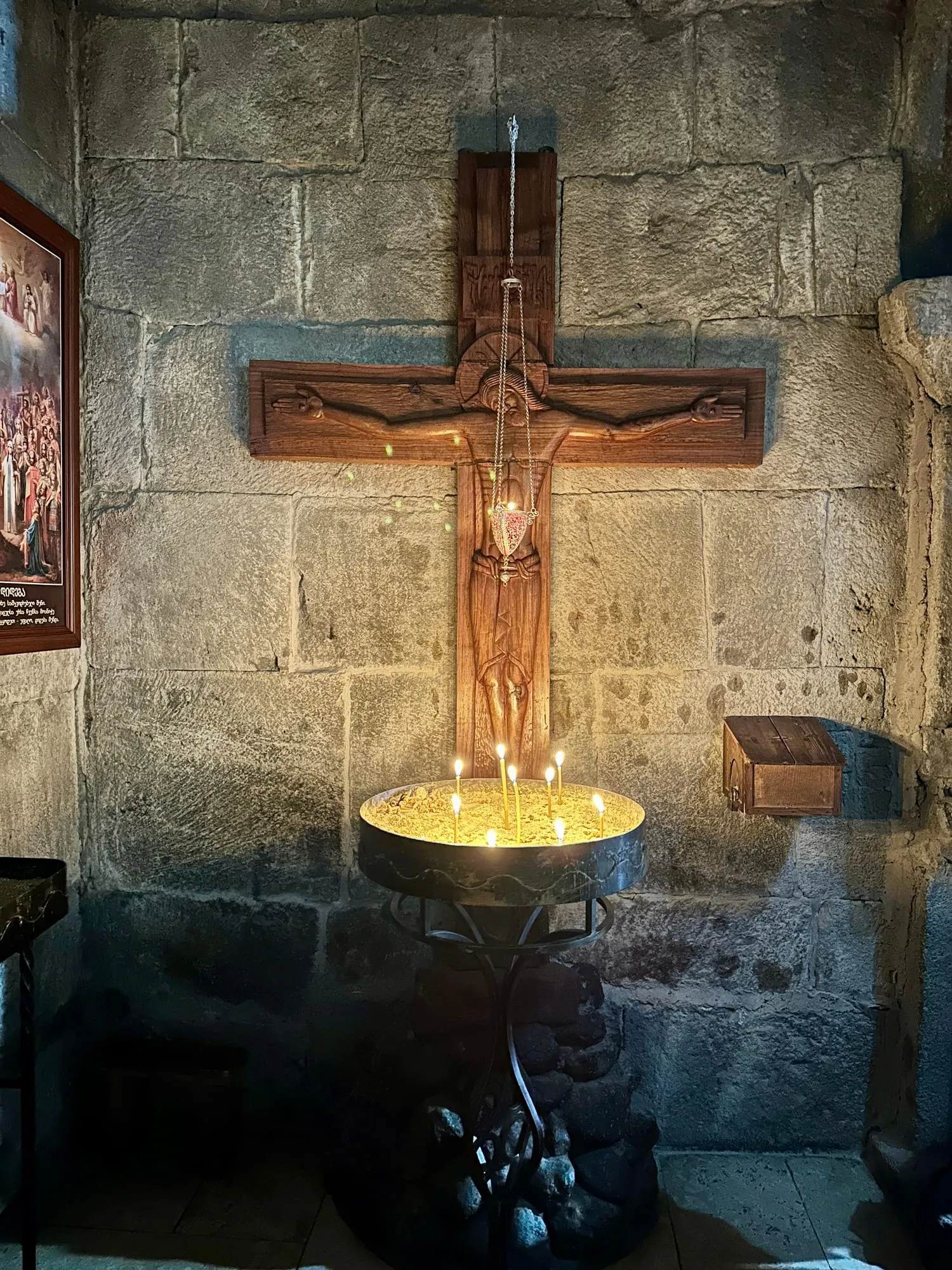 Geschnitztes Holzkreuz mit hängender Lampe und brennenden Kerzen vor einer grob behauenen Steinwand.