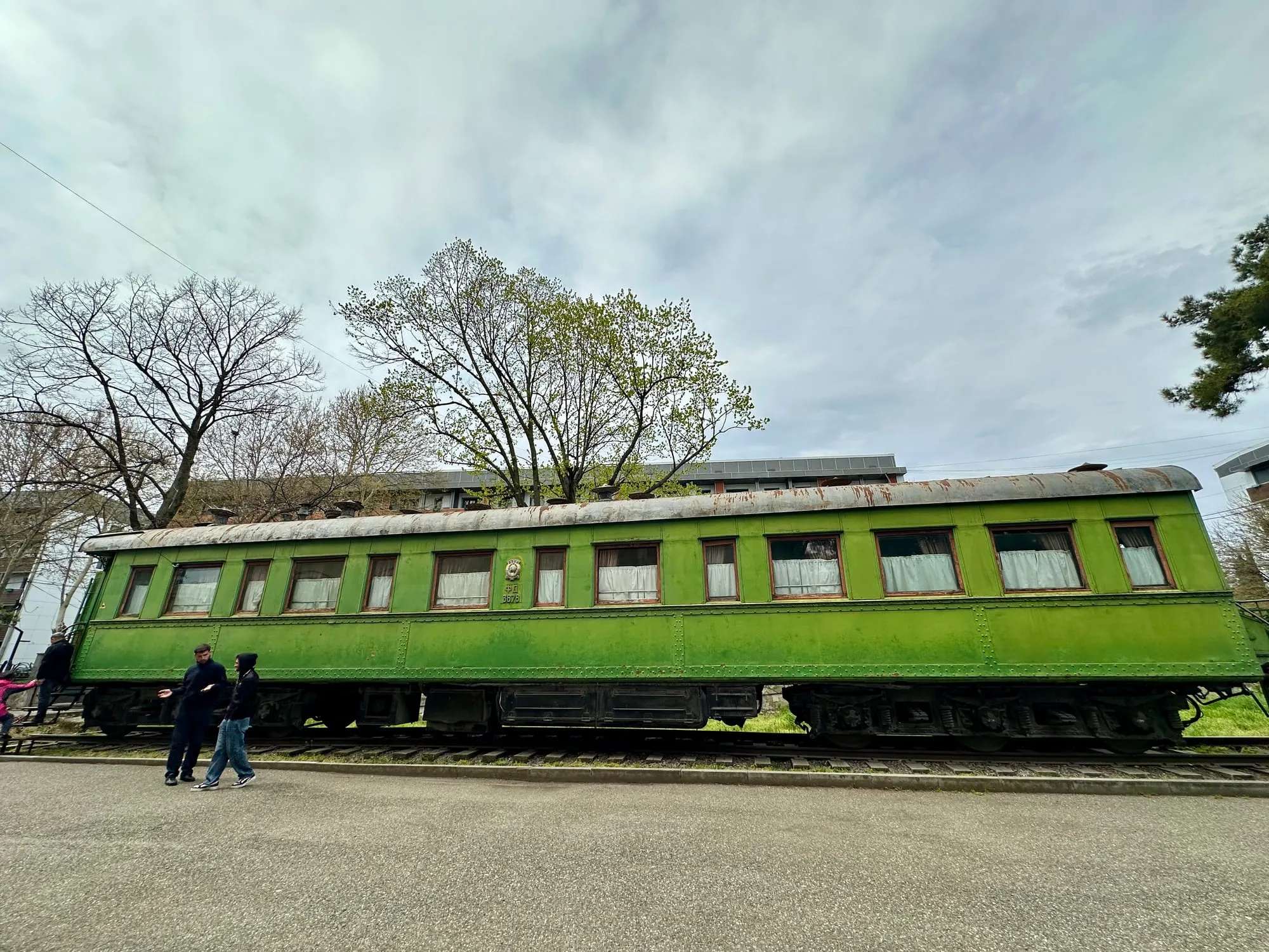 Grüner, stark genieteter Eisenbahnwagen mit rostigem Dach und Fenstern mit Vorhängen steht auf Gleisen, davor mehrere Besucher.