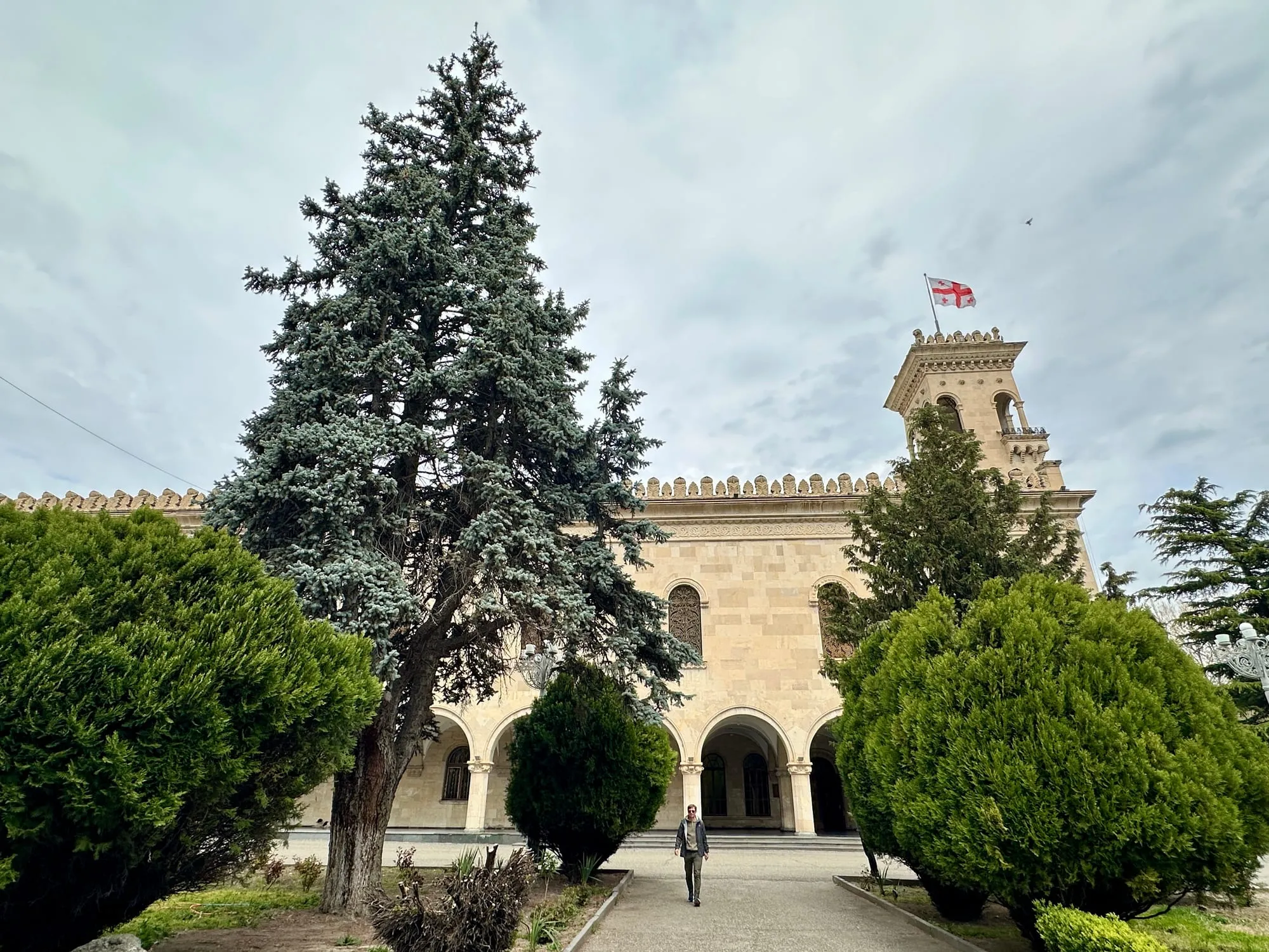 Reich verzierter heller Museumsbau mit Arkaden, teilweise hinter einem hohen Nadelbaum, rechts weht die georgische Flagge.