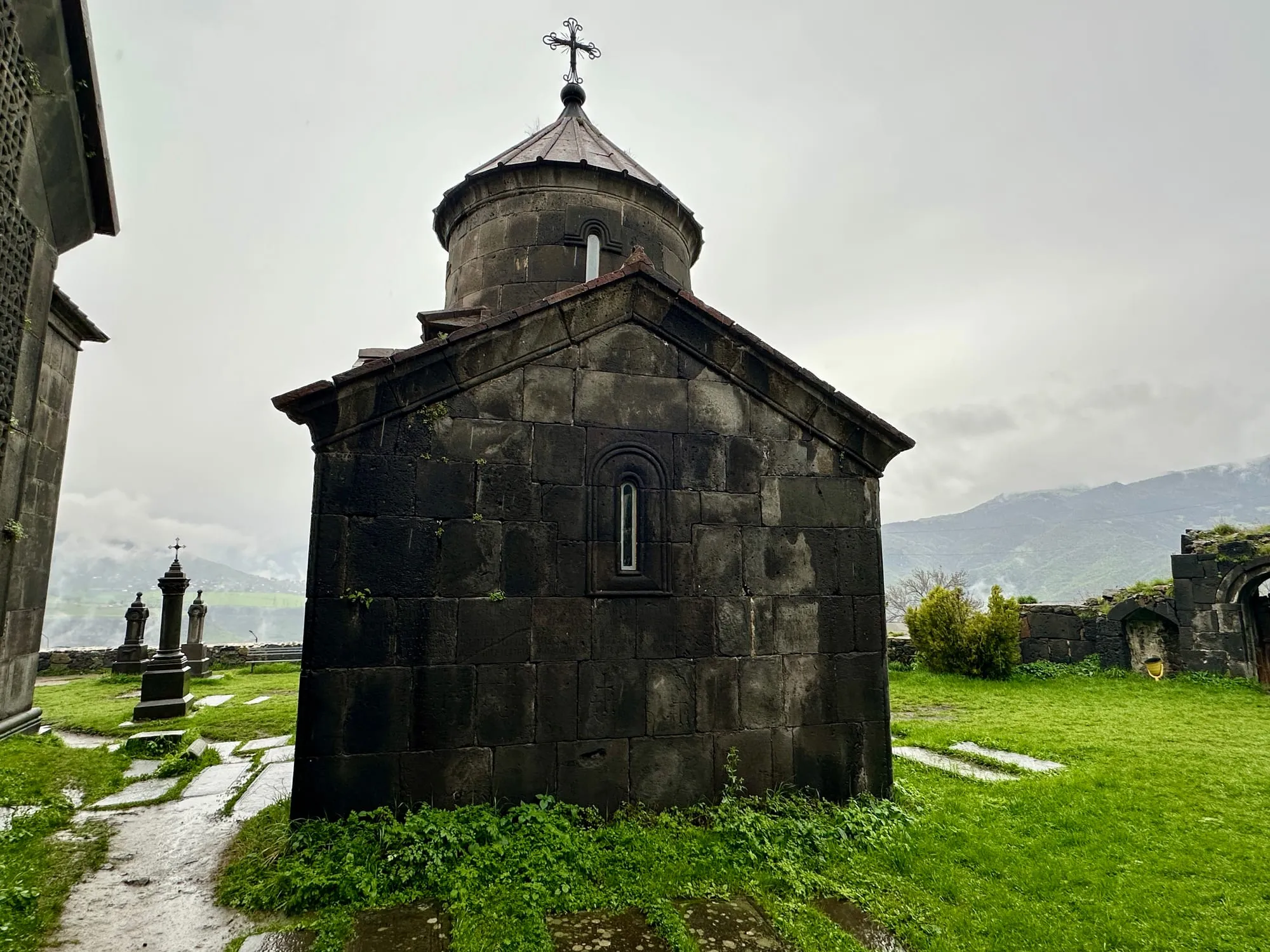 Dunkle Steinkirche mit Kuppel und Kreuz in einem grünen Klosterhof vor wolkenverhangenen Bergen.