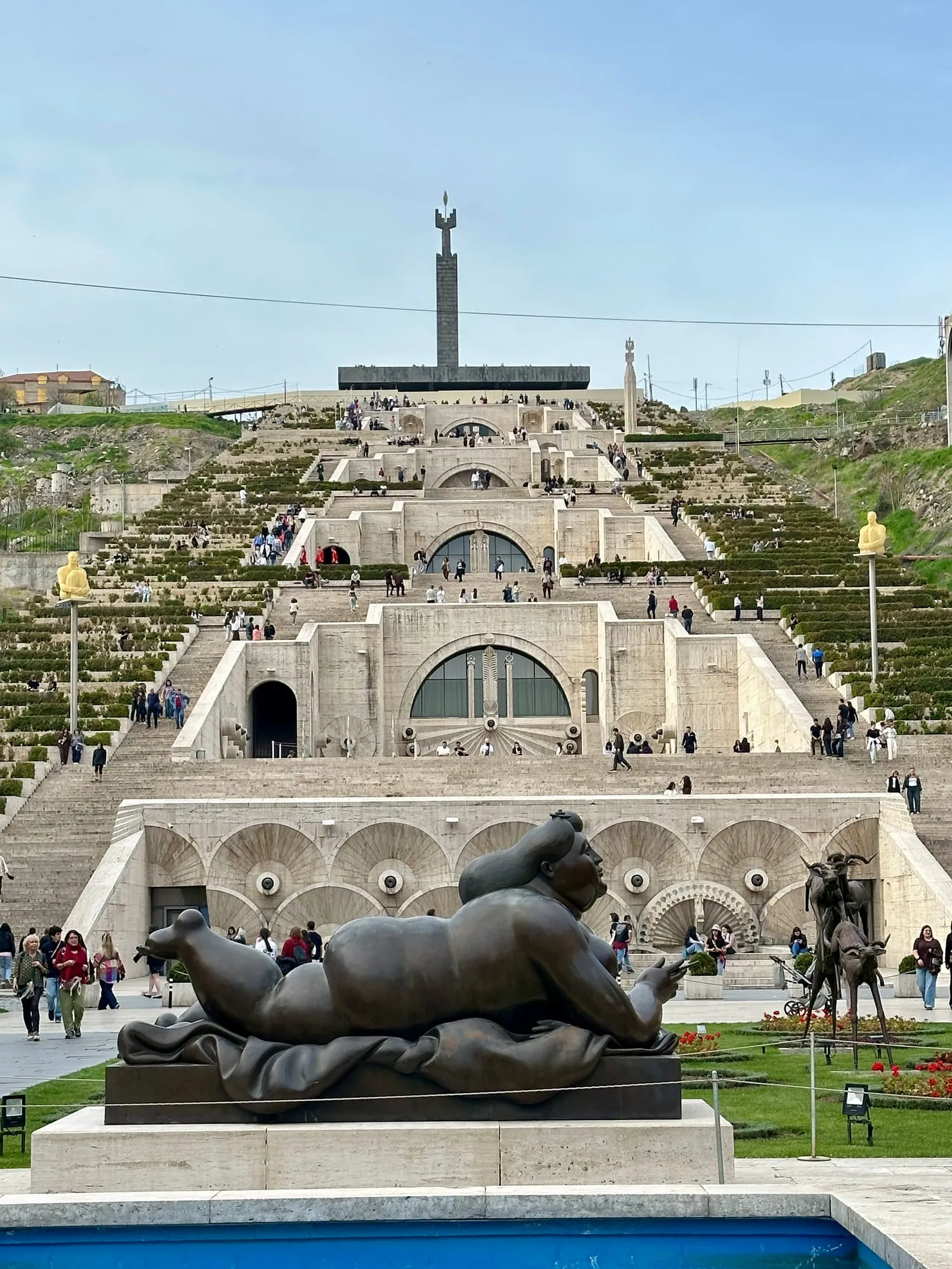 Die monumentale Kaskade in Jerewan mit hellen Steinterrassen, Besuchern, gepflegten Beeten und einer grossen liegenden Tierskulptur im Vordergrund.