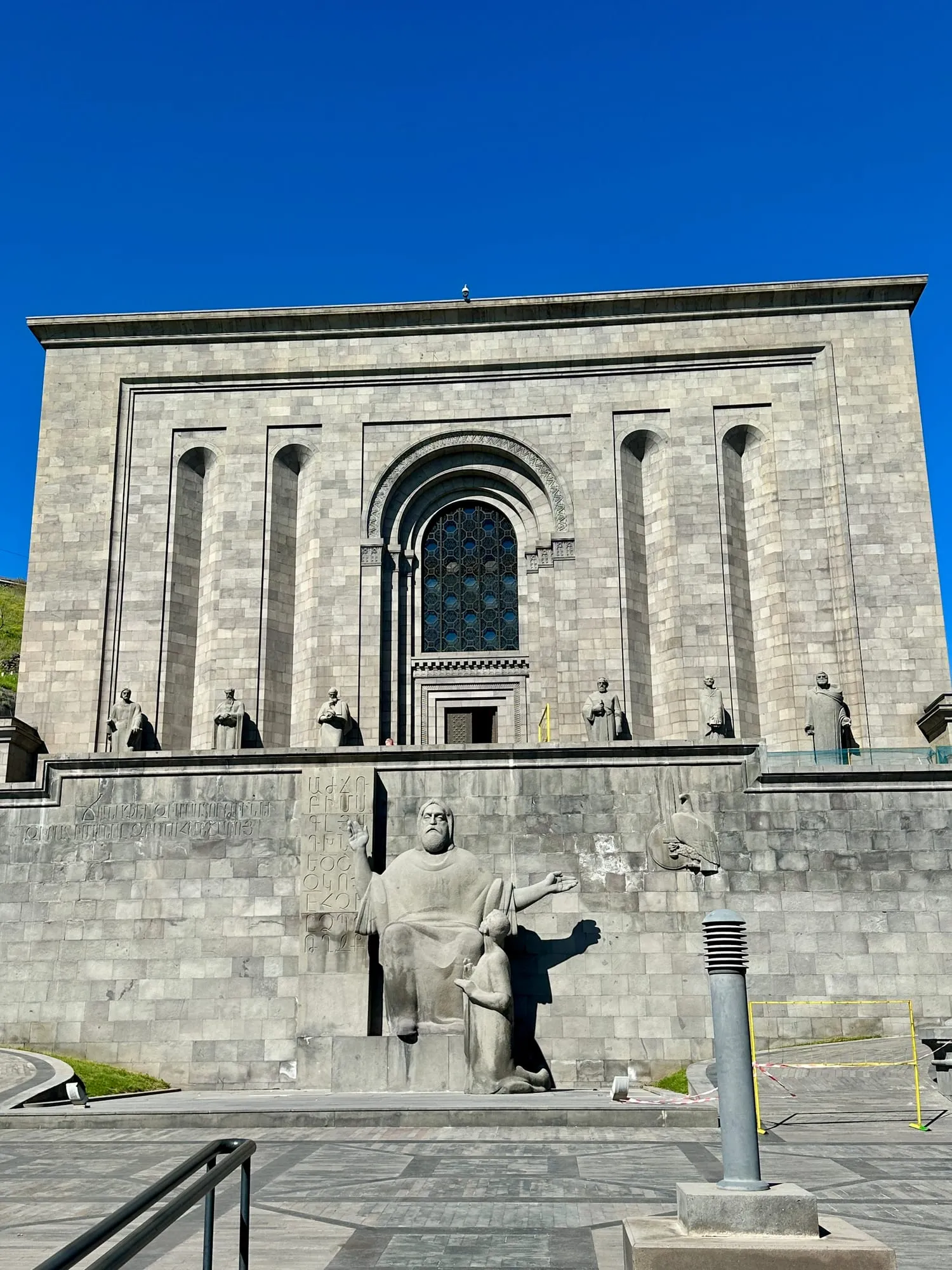 Monumentale Fassade des Matenadaran in Jerewan mit armenischer Inschrift, Rundbogenfenster, steinernen Figuren und einer sitzenden Statue.