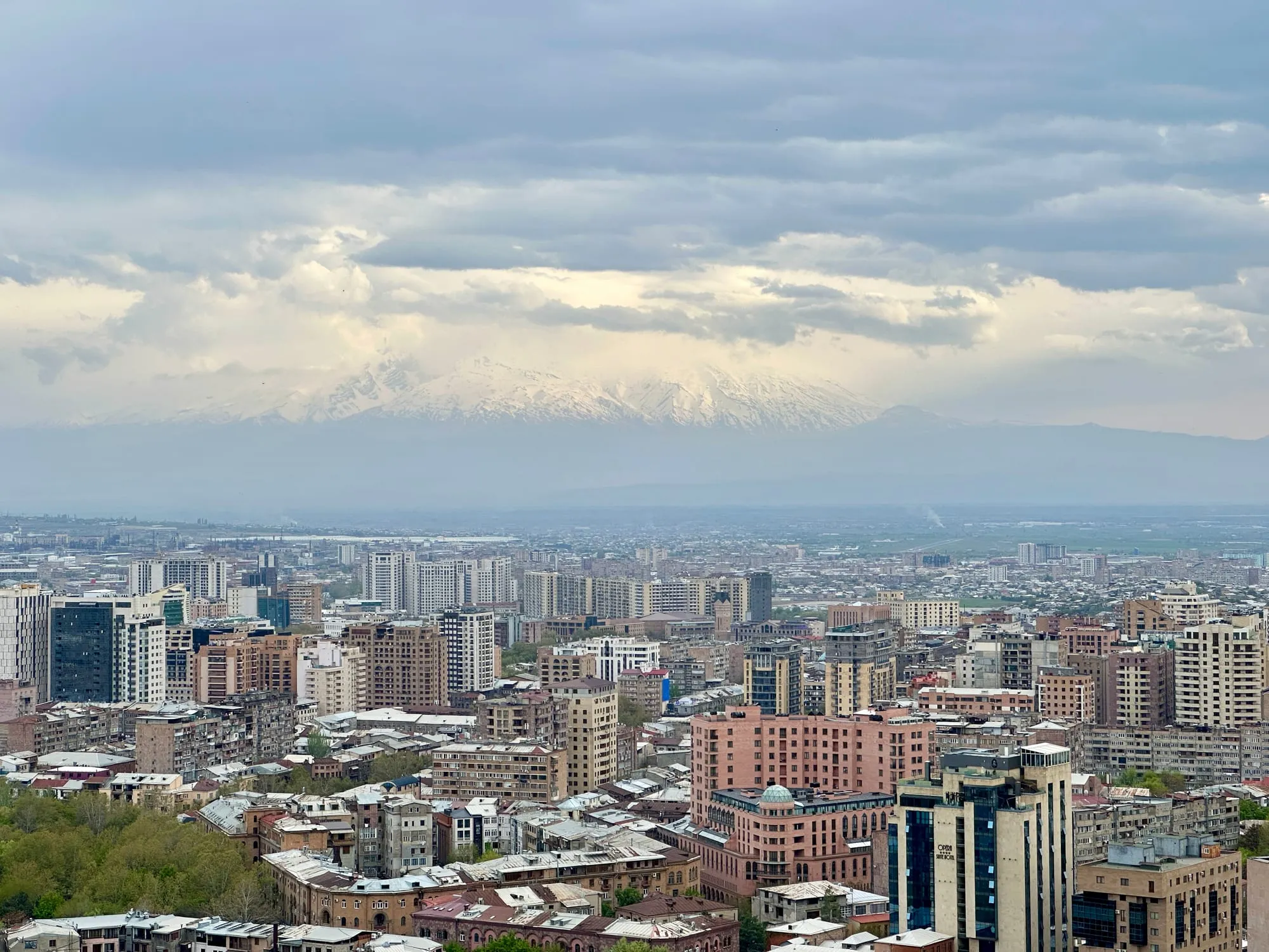 Panorama über Jerewan mit dicht bebauter Innenstadt und einem teilweise von Wolken verhüllten, schneebedeckten Bergmassiv im Hintergrund.