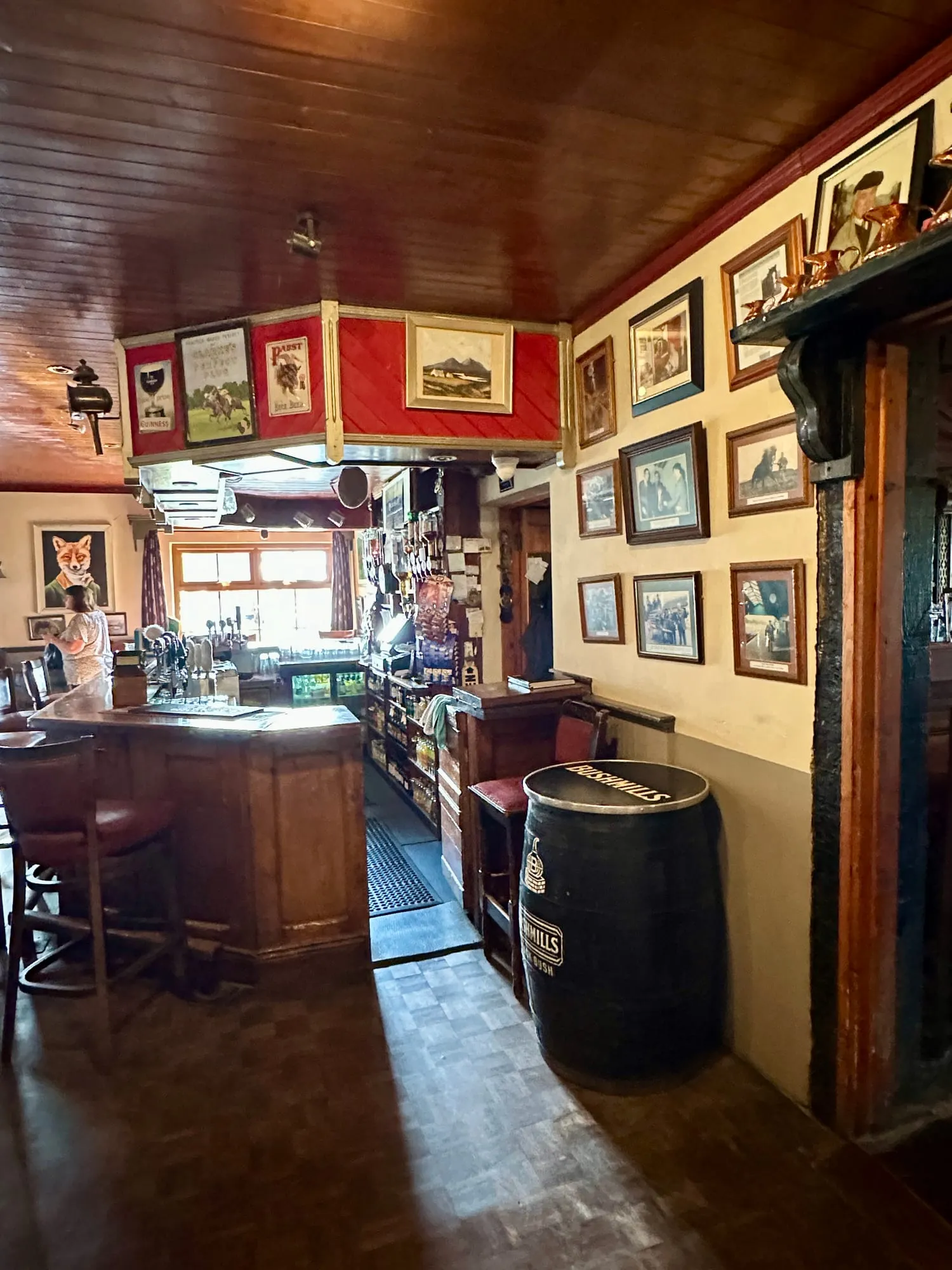 Innenraum eines irischen Pubs mit dunklem Holztresen, Whiskeyfass und dicht behängten Wänden.