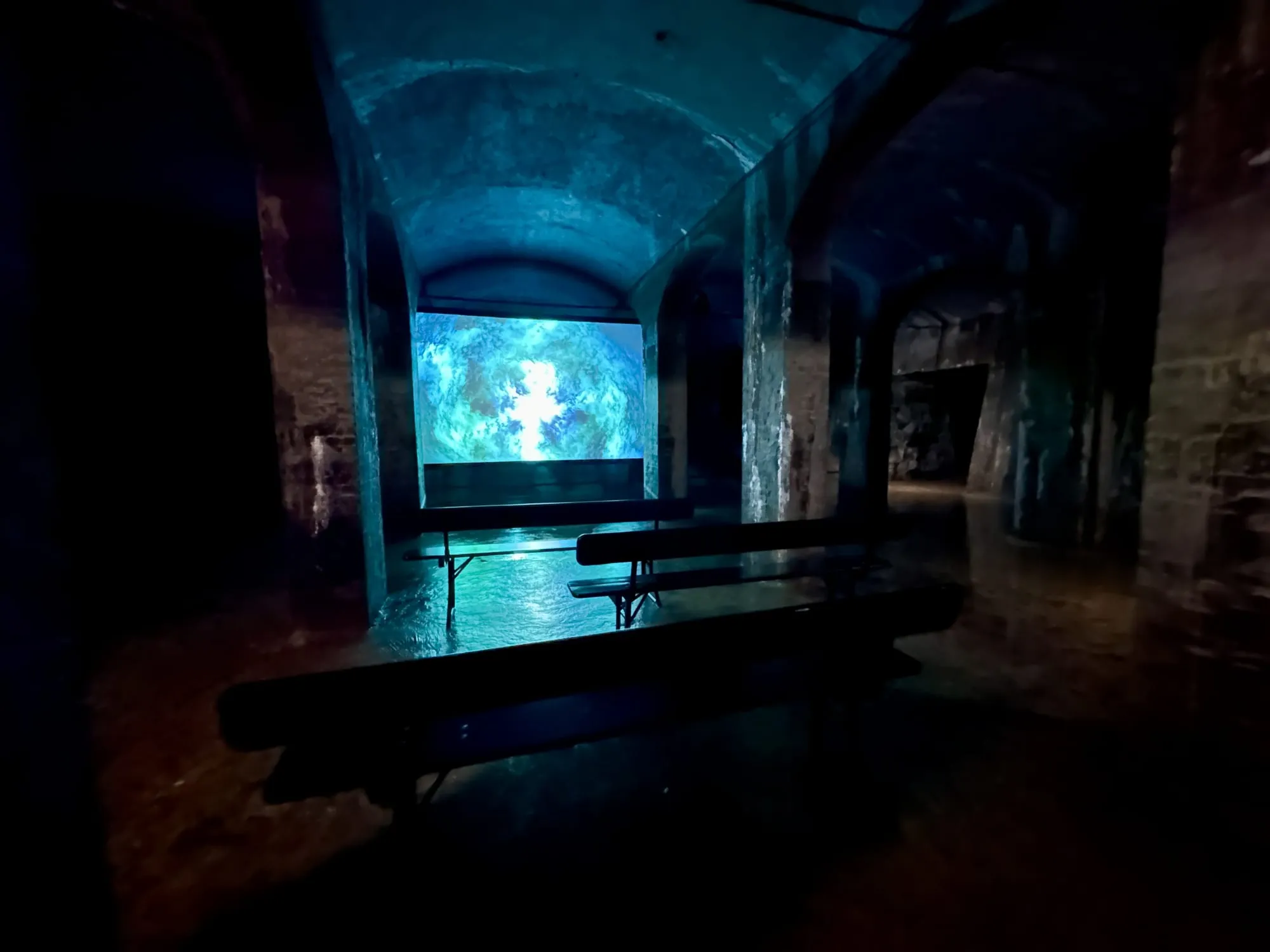 Blau beleuchtetes Gewölbe der Cisternerne mit Videoleinwand und Sitzbänken auf nassem Boden.