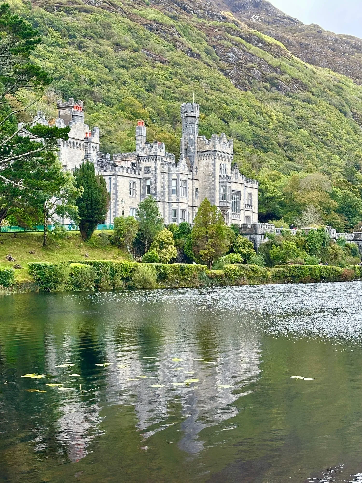 Kylemore Abbey aus grauem Granit am bewaldeten Hang, die Fassade spiegelt sich im See davor.