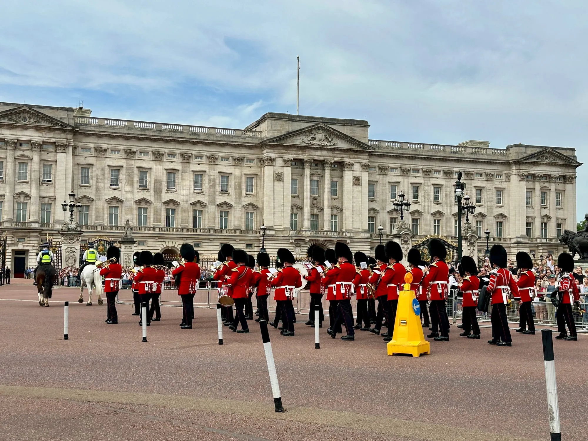 Musikkorps und Gardisten in roten Uniformen mit Bärenfellmützen vor dem Buckingham Palace, dahinter Zuschauer hinter Absperrgittern.