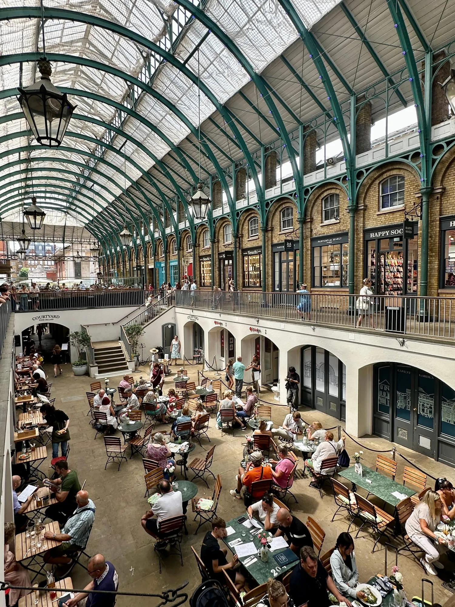 Innenraum der Markthalle von Covent Garden mit gläsernem Tonnendach, grüner Eisenkonstruktion und Cafétischen im tiefergelegten Hof.
