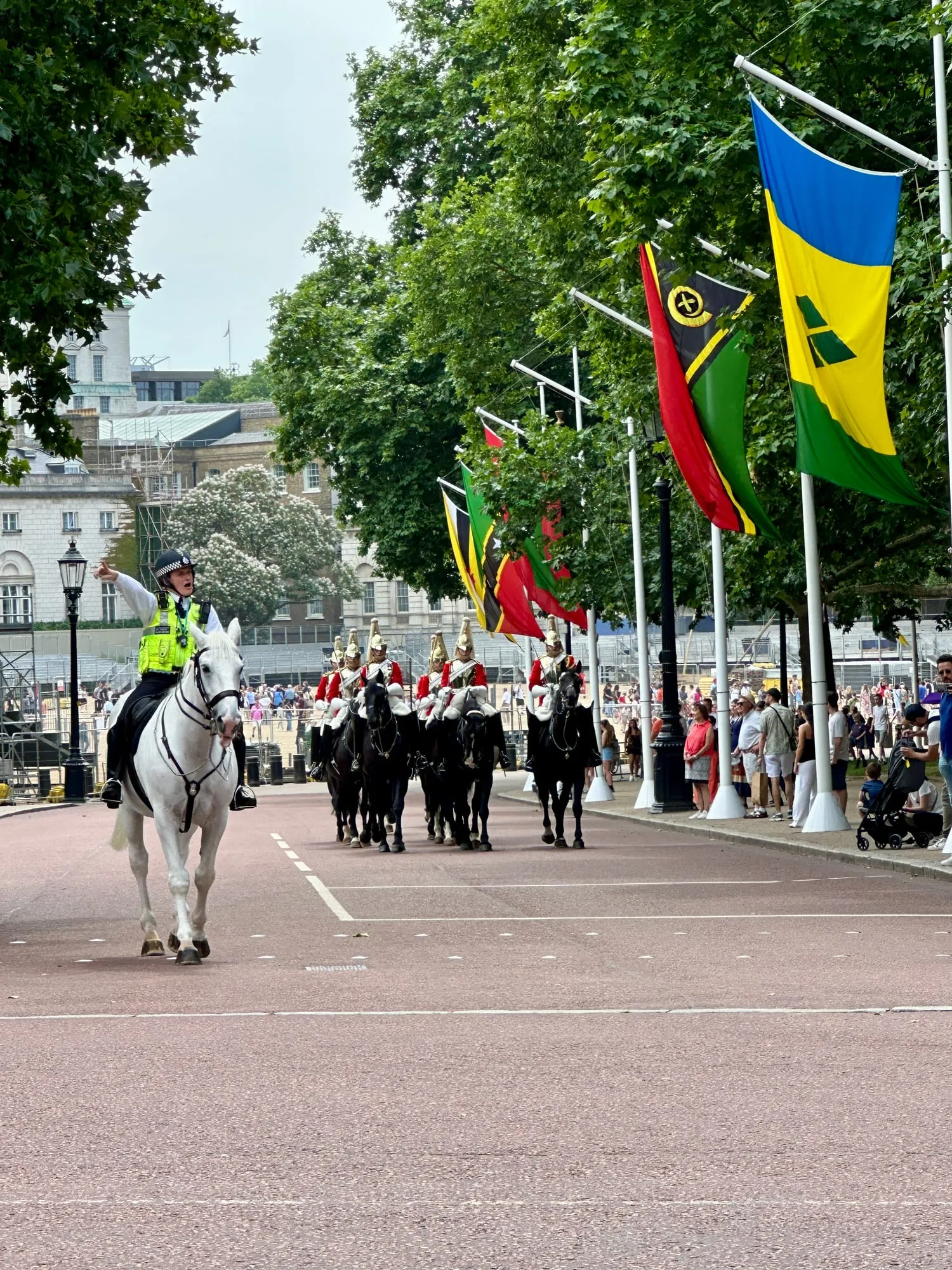 Berittene Soldaten der Household Cavalry mit Helmbusch reiten die Prachtstrasse The Mall hinunter, begleitet von einer Polizeireiterin.