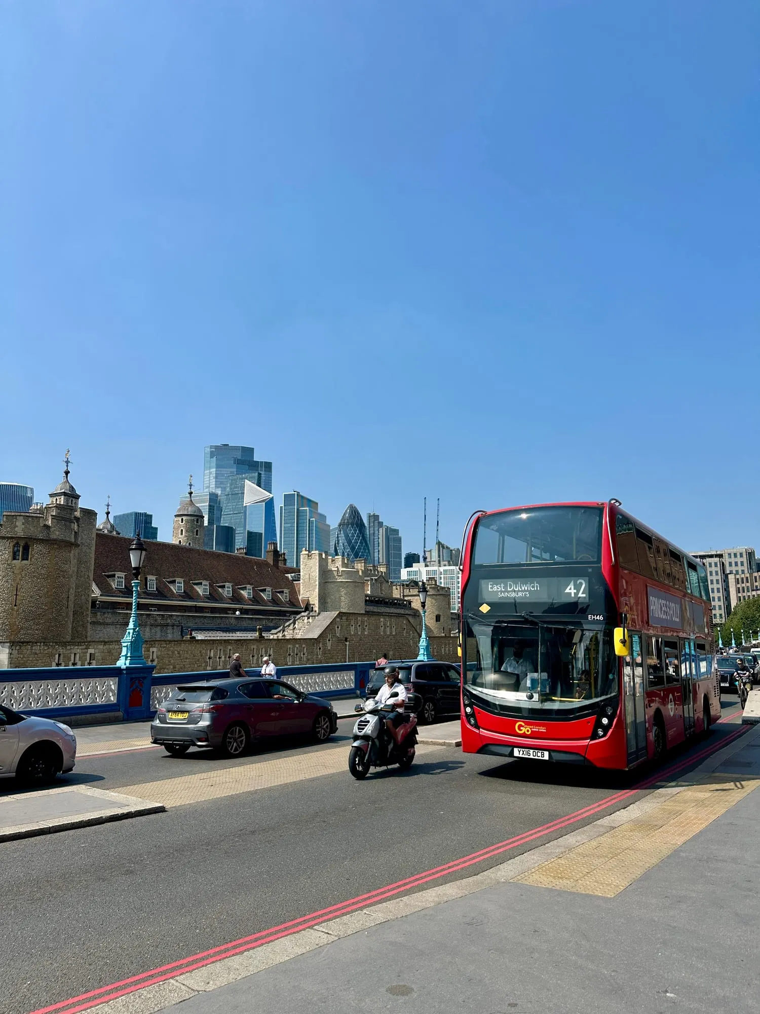 Roter Doppeldeckerbus auf der Tower Bridge, dahinter der Tower of London und die Hochhäuser der City.
