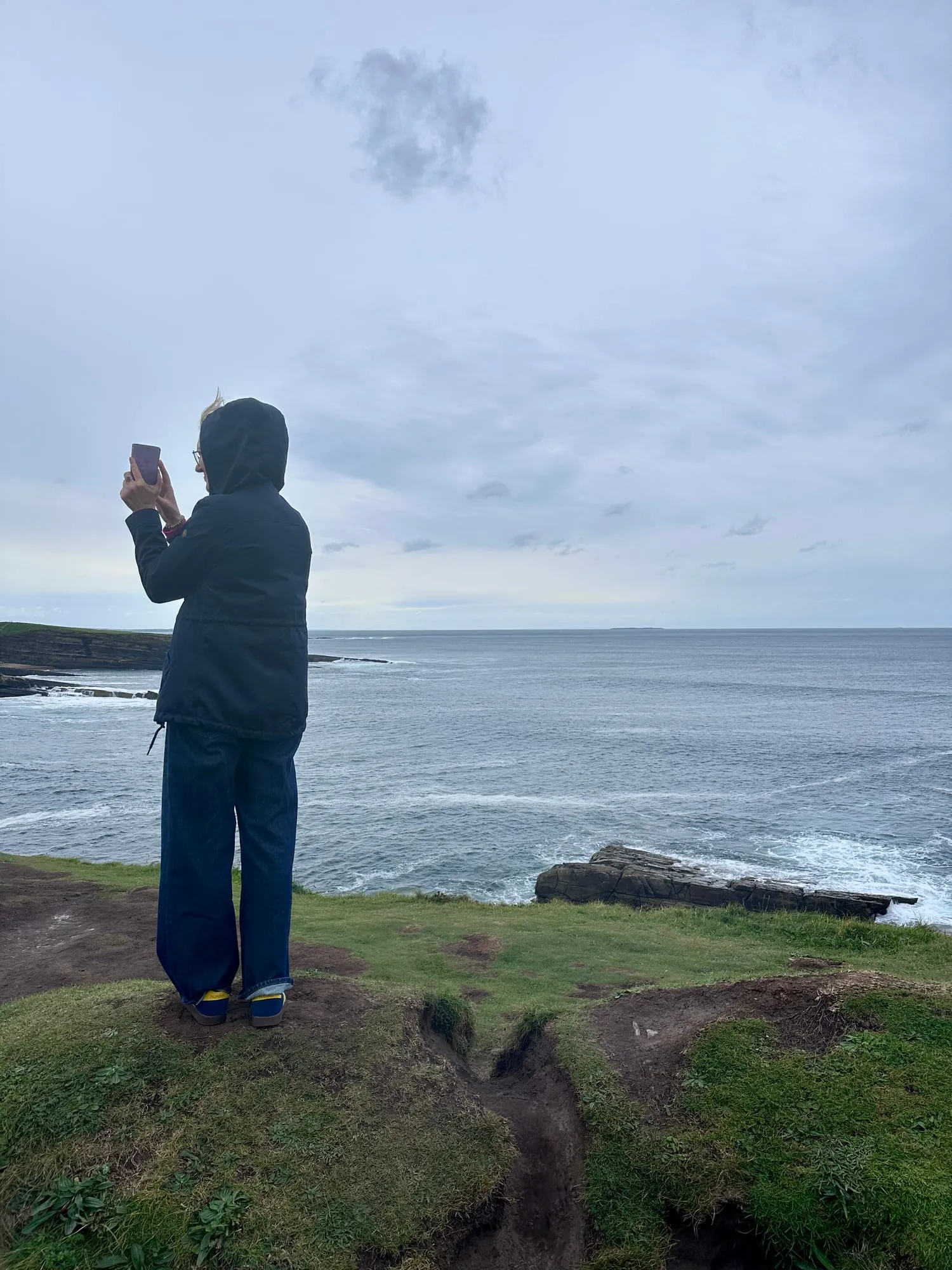 Person in Regenjacke steht auf der Klippe von Mullaghmore und fotografiert den Atlantik.