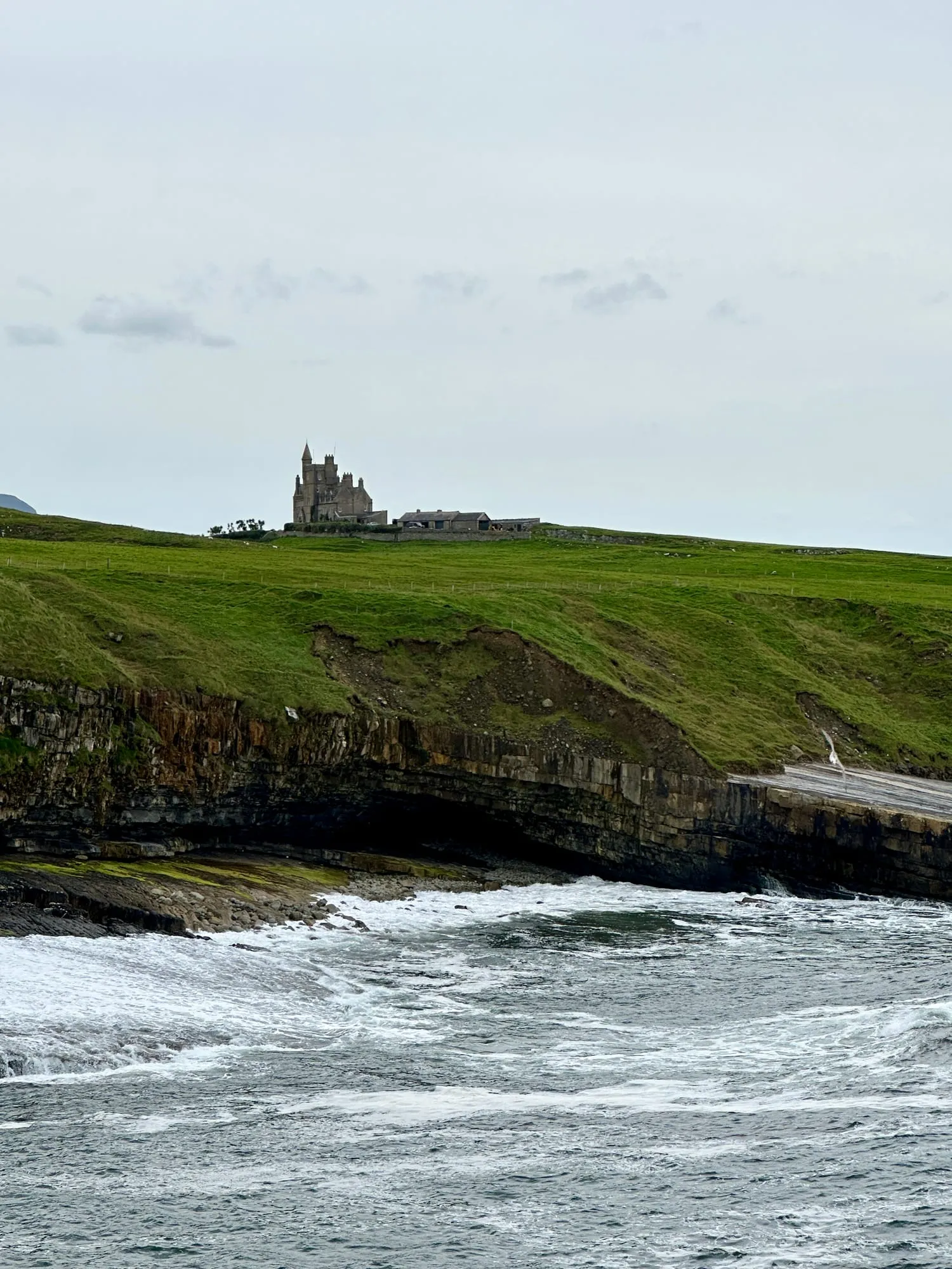 Classiebawn Castle auf der Klippe über dem Meer, davor grüne Weiden und die Brandung der Bucht.