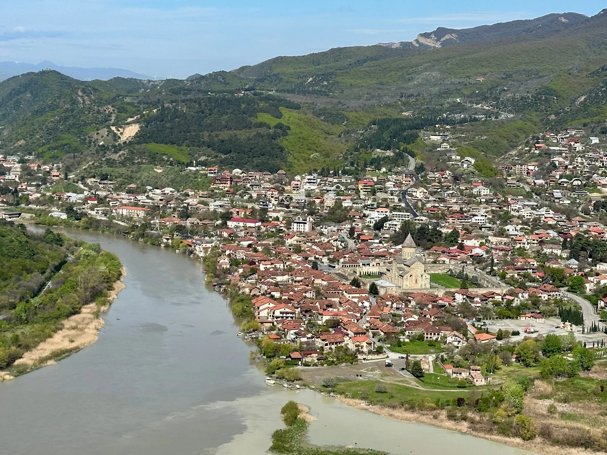 Panoramablick vom Dschwari-Kloster auf Mzcheta zwischen bewaldeten Hügeln, die Stadt liegt am Zusammenfluss zweier Flüsse, rechts unten eine grosse steinerne Kirche.