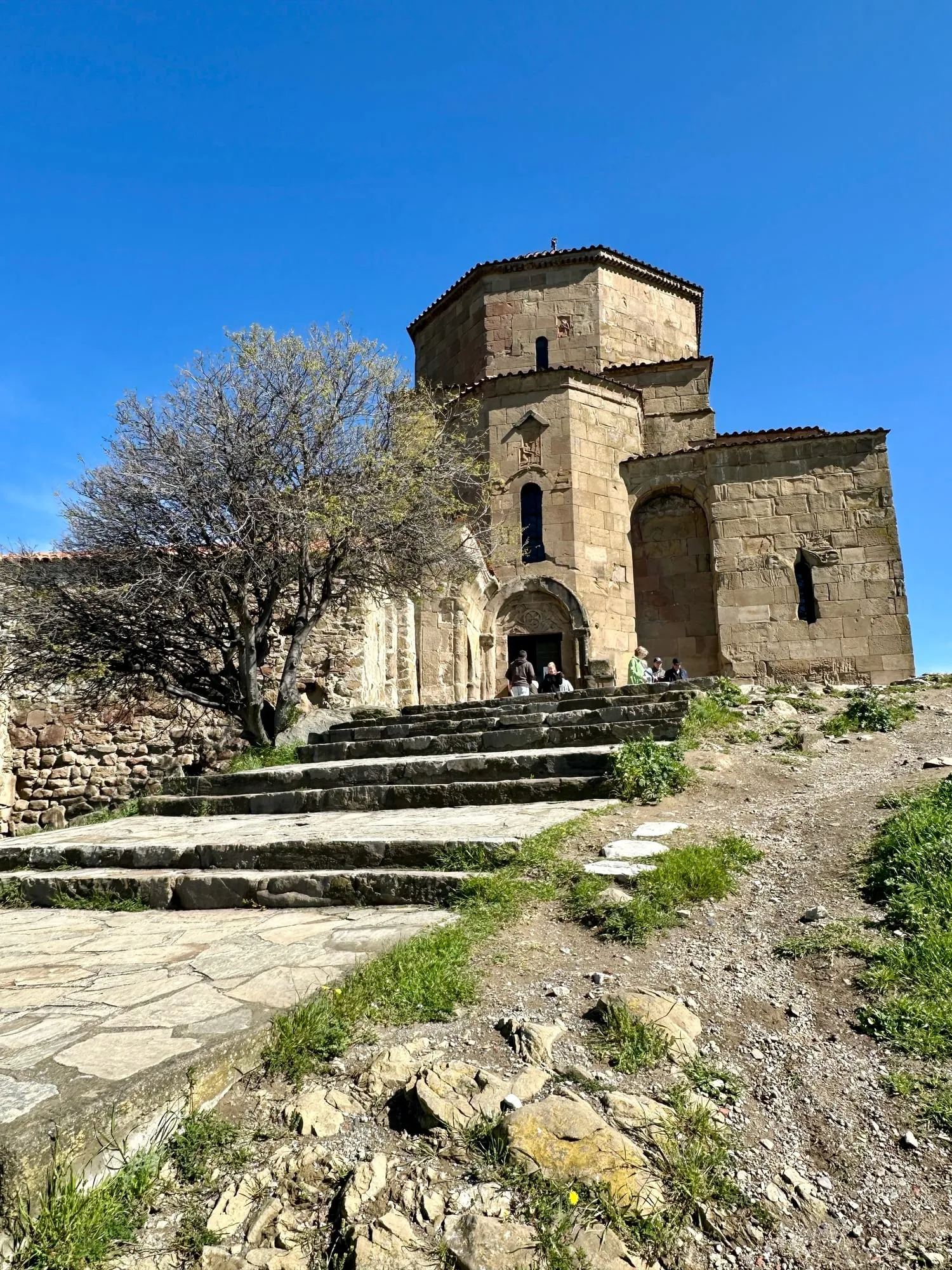 Steinerne Kirche des Dschwari-Klosters mit reliefgeschmücktem Portal, breiter Treppe und Besuchern unter blauem Himmel.