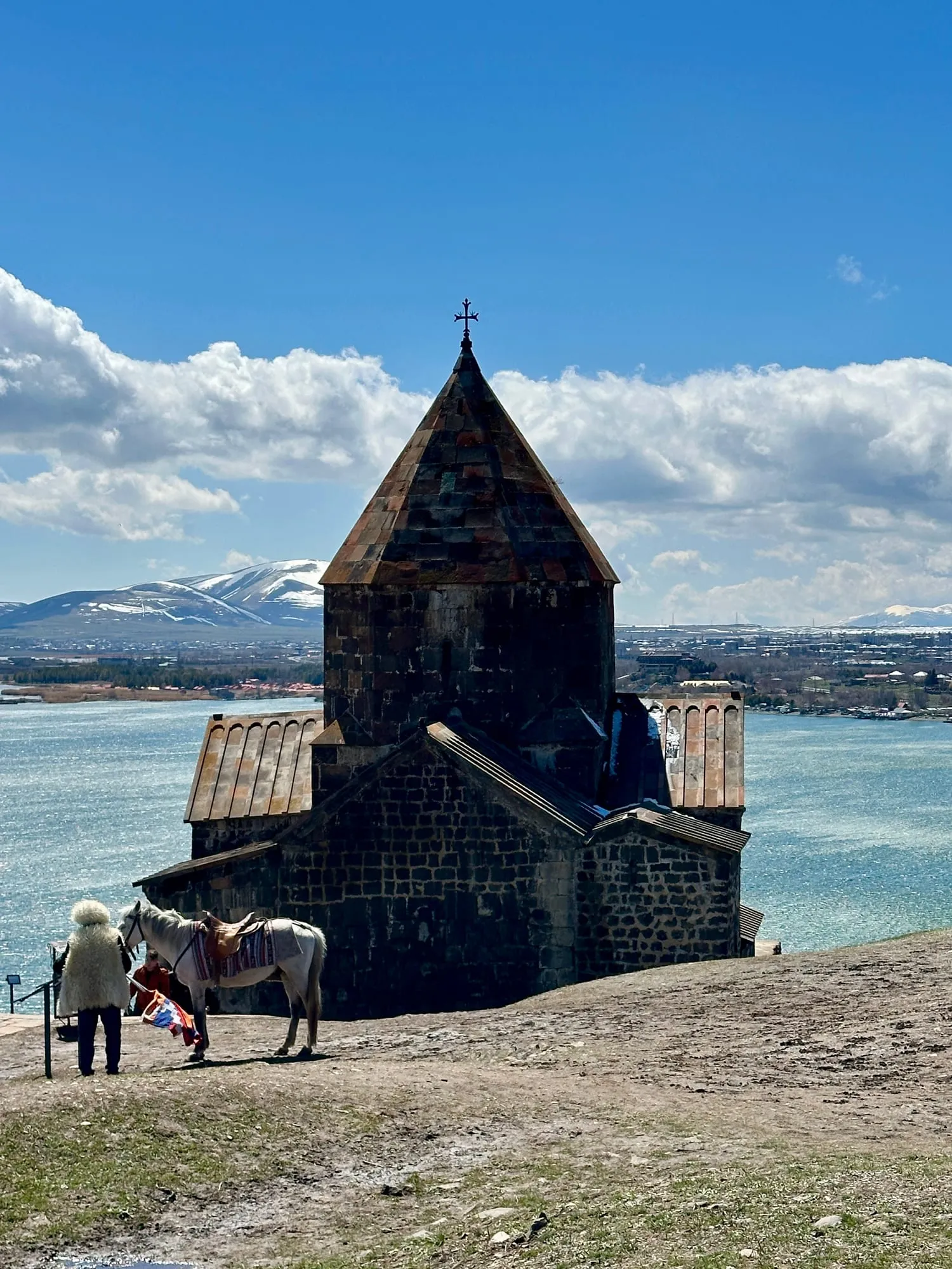 Dunkle Steinkirche mit Kreuz am Sewansee, dahinter schneebedeckte Berge, im Vordergrund ein weisses Pferd neben einer Person.