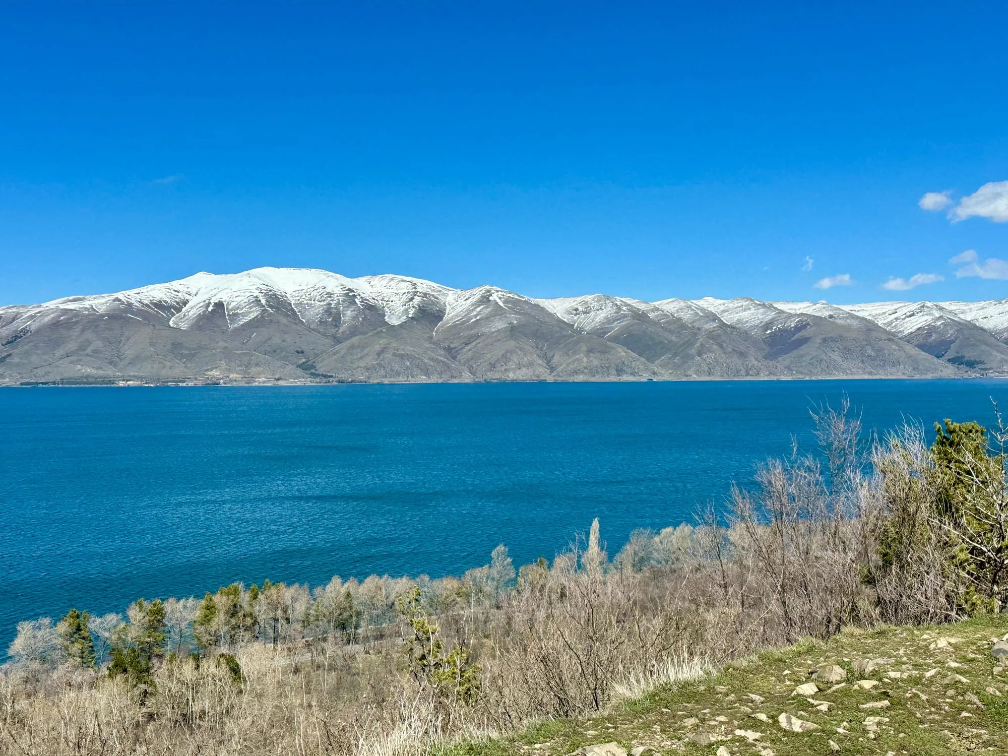Tiefblauer Sewansee vor schneebedeckten Bergen, mit kahlem Buschland und einem steinigen Hang im Vordergrund.