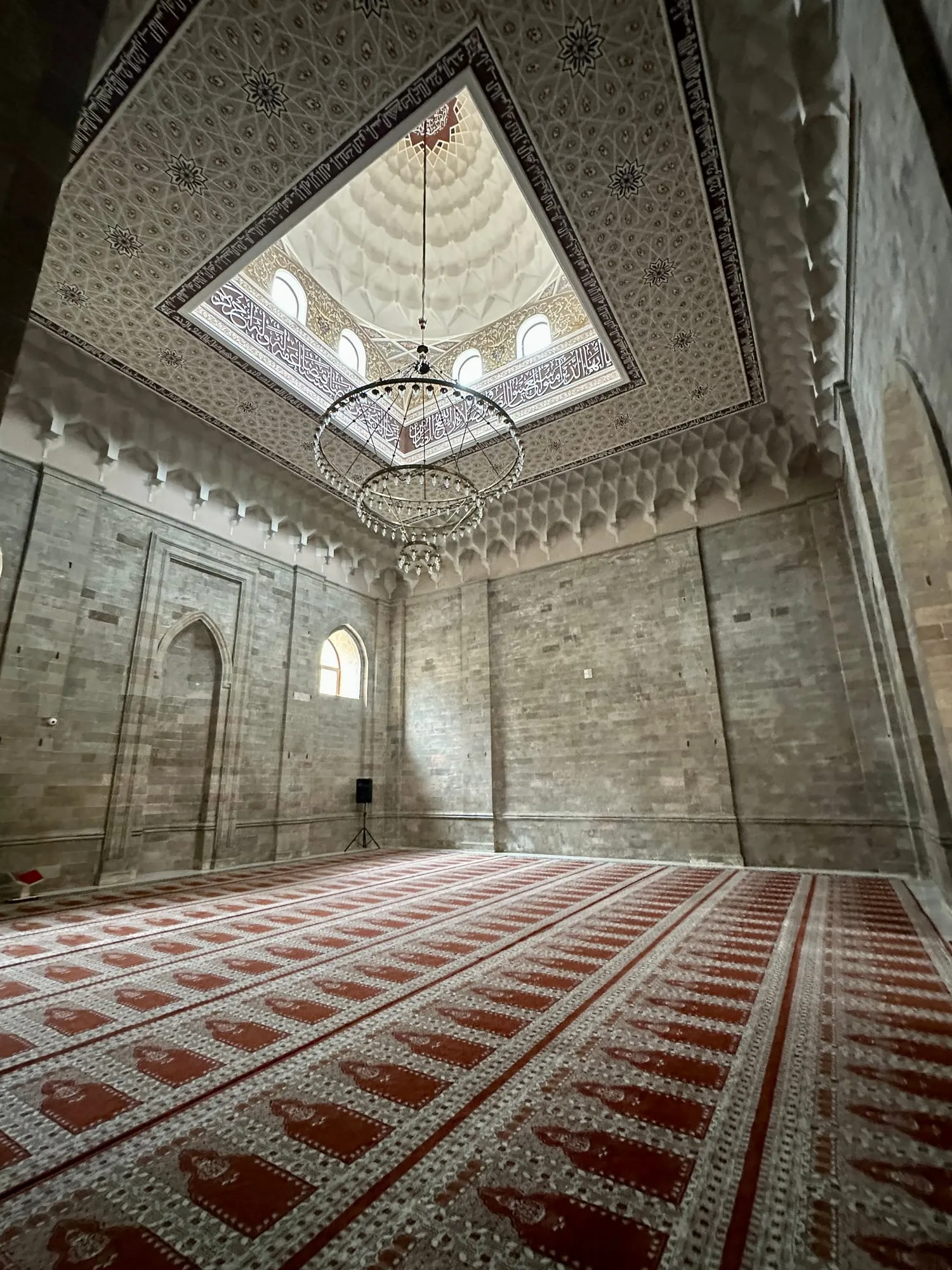 Reich verzierter Moscheeraum mit arabischer Kalligrafie an der Decke, Kronleuchtern und rot gemustertem Gebetsteppich.