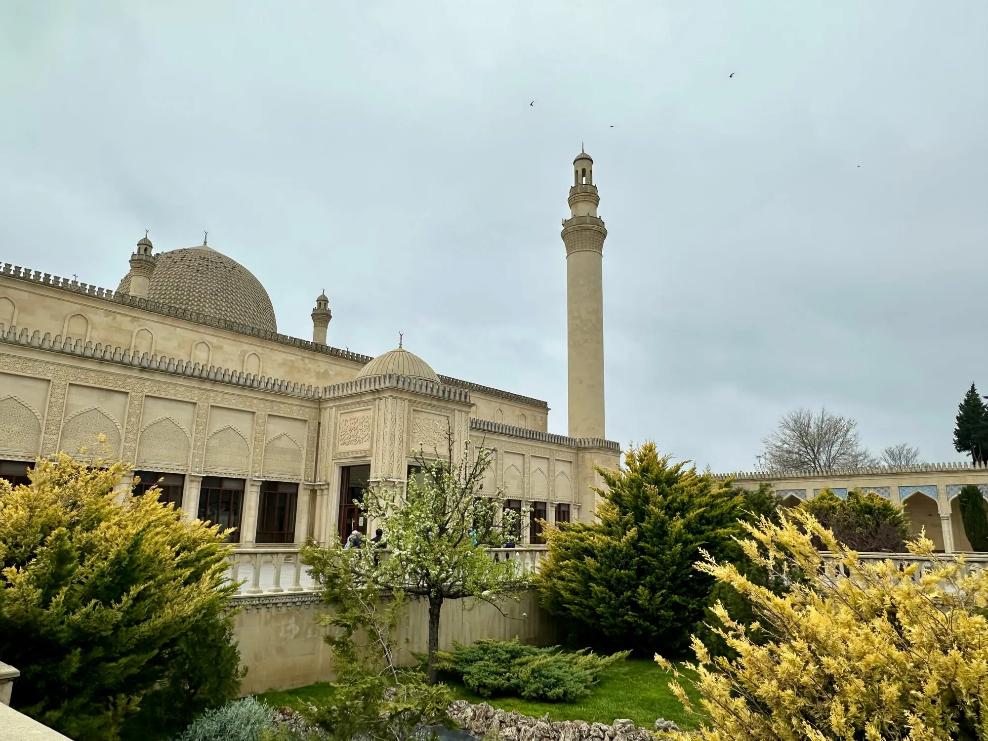 Helle Moschee mit mehreren Kuppeln und einem hohen Minarett, umgeben von dichtem Grün unter hellem Himmel.