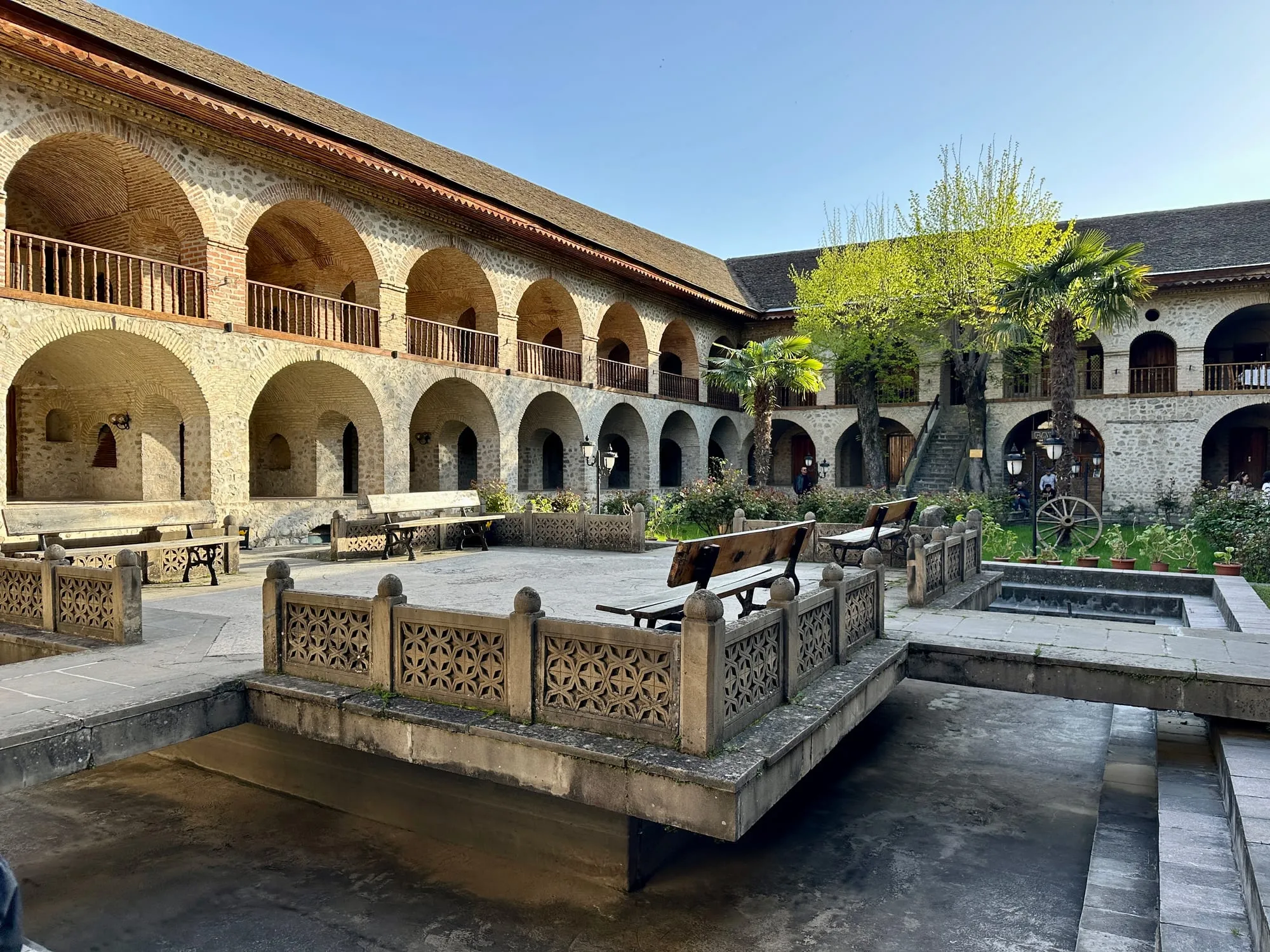 Innenhof der Karawanserei von Şəki mit zweigeschossigen Steinarkaden, Bänken und Palmen.