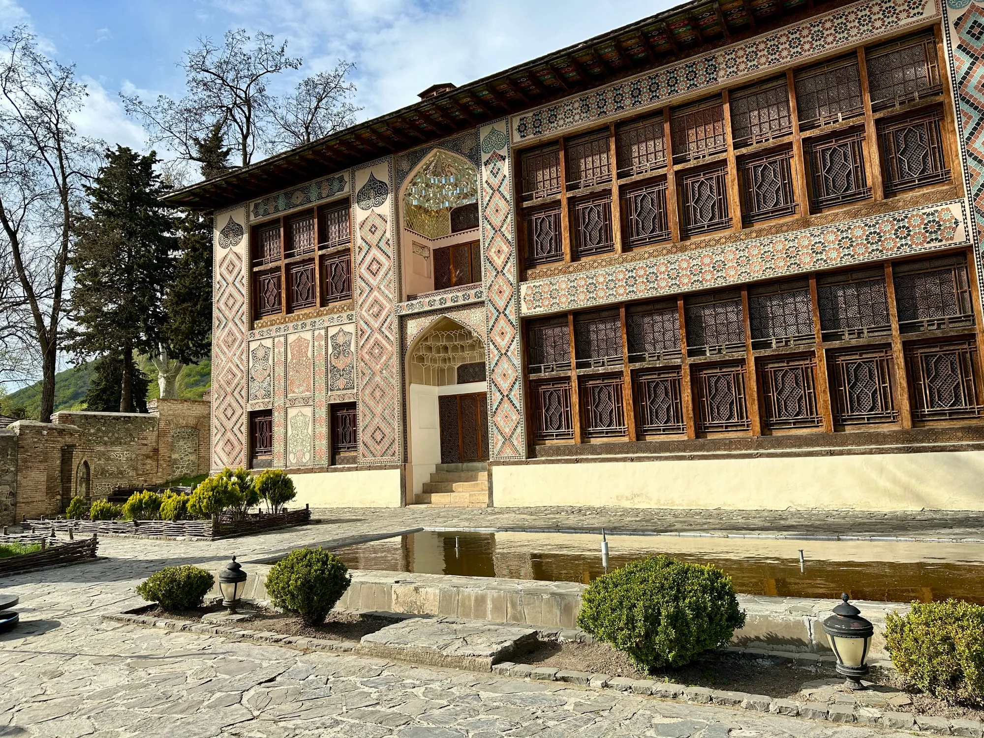 Fassade des Palasts der Schaki-Khane mit Fresken, farbigen Mosaiken, vergitterten Şəbəkə-Fenstern und begrüntem Innenhof.