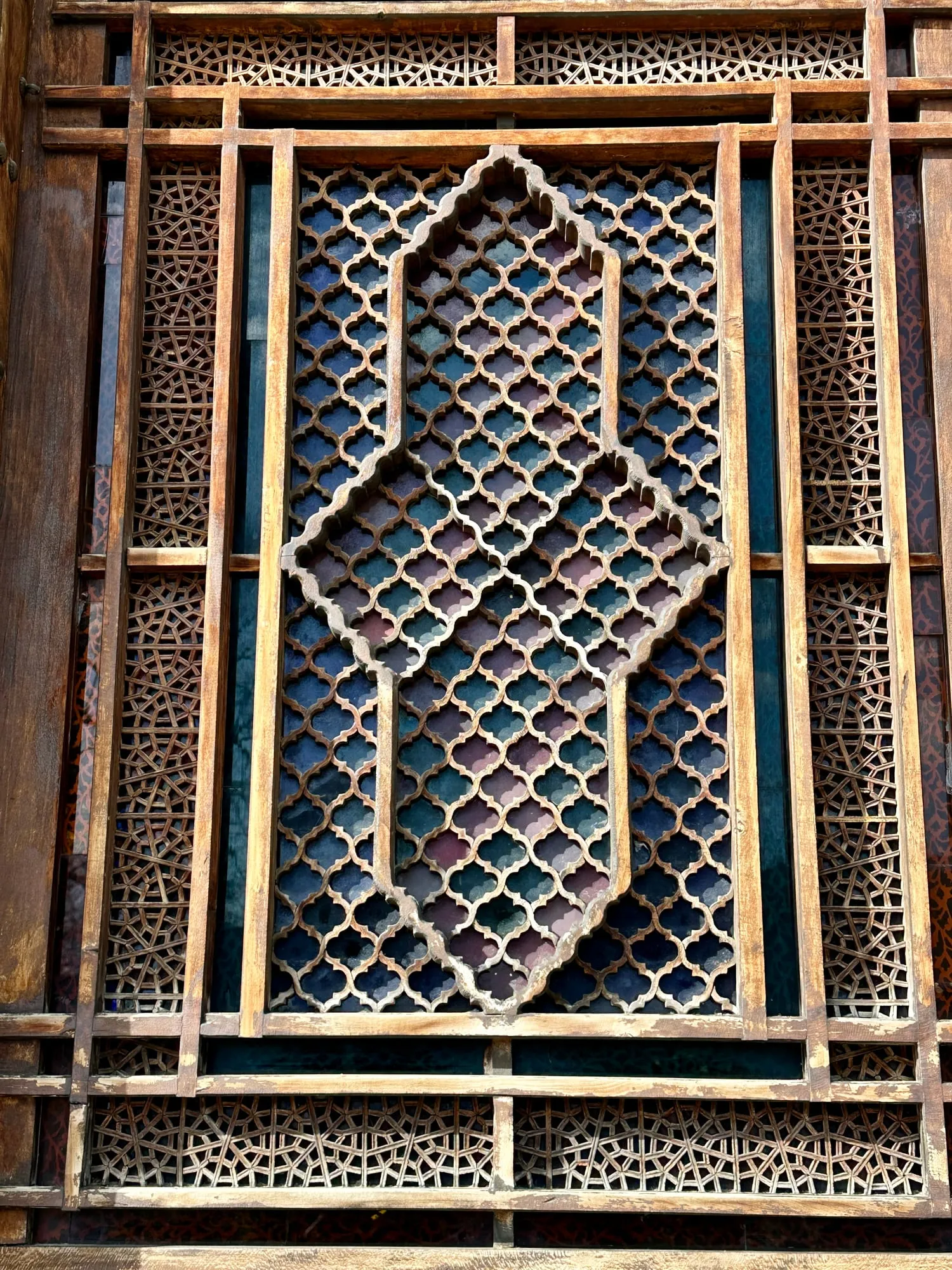 Nahaufnahme eines Şəbəkə-Fensters mit geometrischem Holzgitter und blauvioletten Glasfeldern.