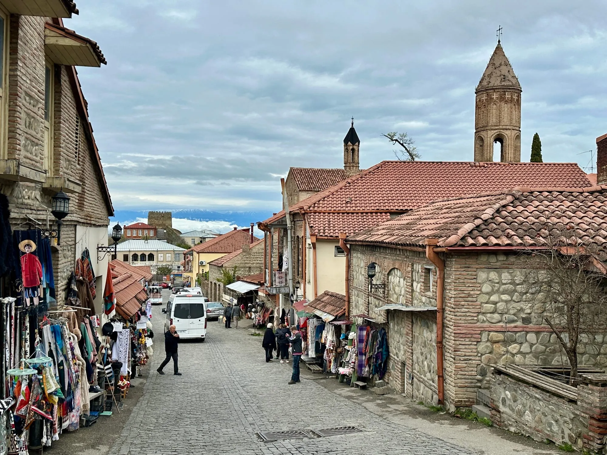 Kopfsteinpflastergasse in der Altstadt von Signagi mit roten Ziegeldächern, Souvenirständen, Türmen und schneebedeckten Bergen im Hintergrund.