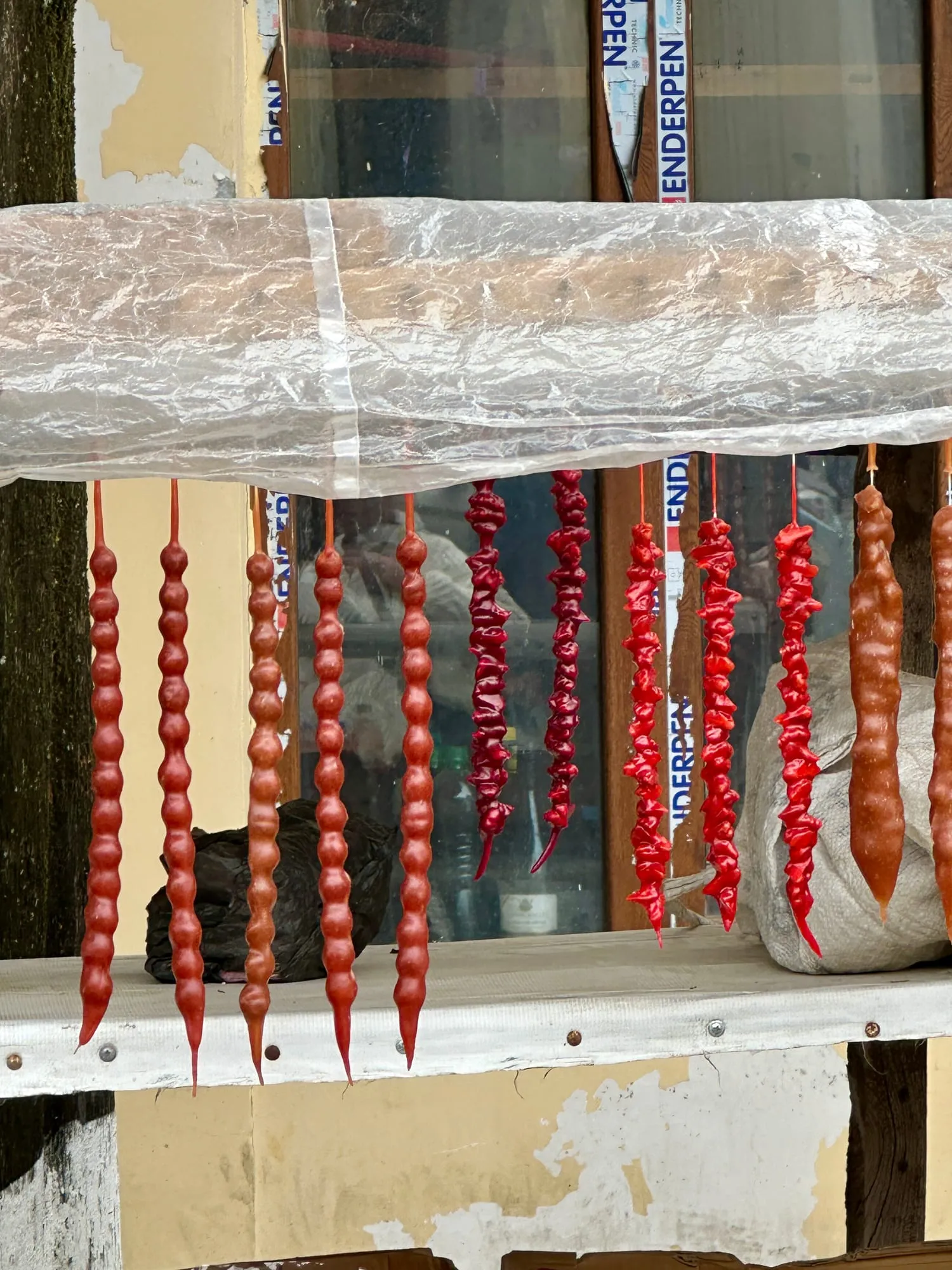 Lange, knubbelige braune Tschurtschchela und weitere rote getrocknete Waren hängen vor einem Marktfenster.
