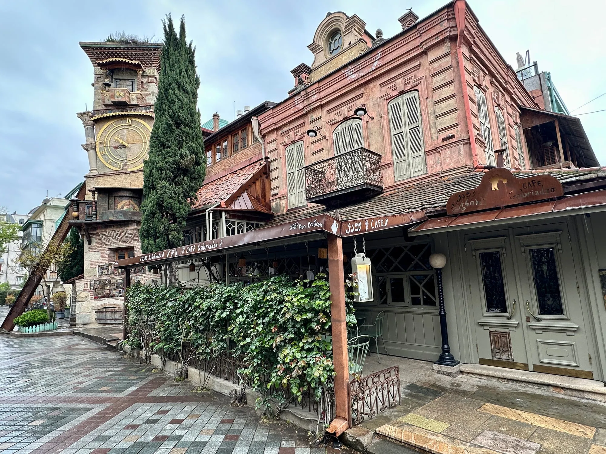 Ornamentierter Uhrturm des Rezo-Gabriadze-Marionettentheaters neben einem rosafarbenen Altstadthaus mit Café in Tbilisi.