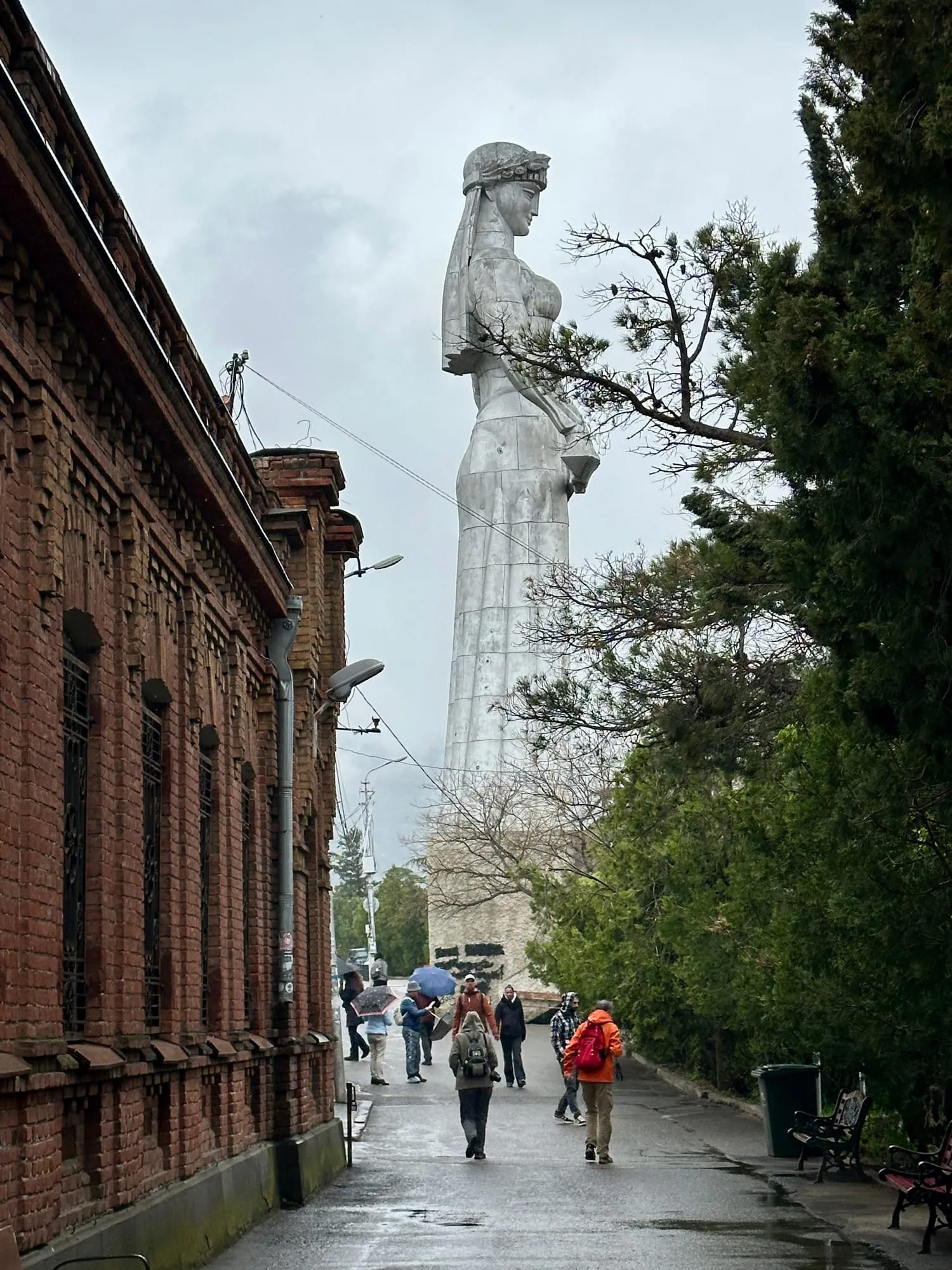Die monumentale graue Frauenstatue Kartlis Deda über einem nassen Weg zwischen Backsteinmauer und Bäumen, mehrere Menschen tragen Regenschirme.