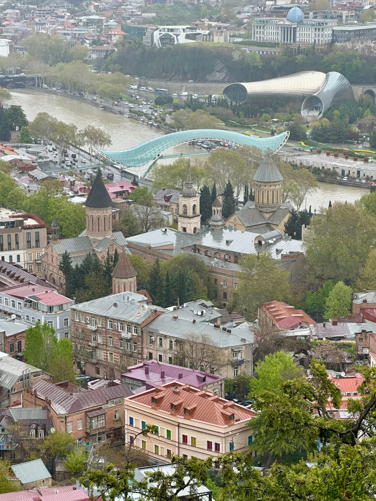 Panoramablick über Tbilisi mit der Kura, der gläsernen Friedensbrücke, dicht bebauten Stadthängen und Kirchtürmen im Vordergrund.