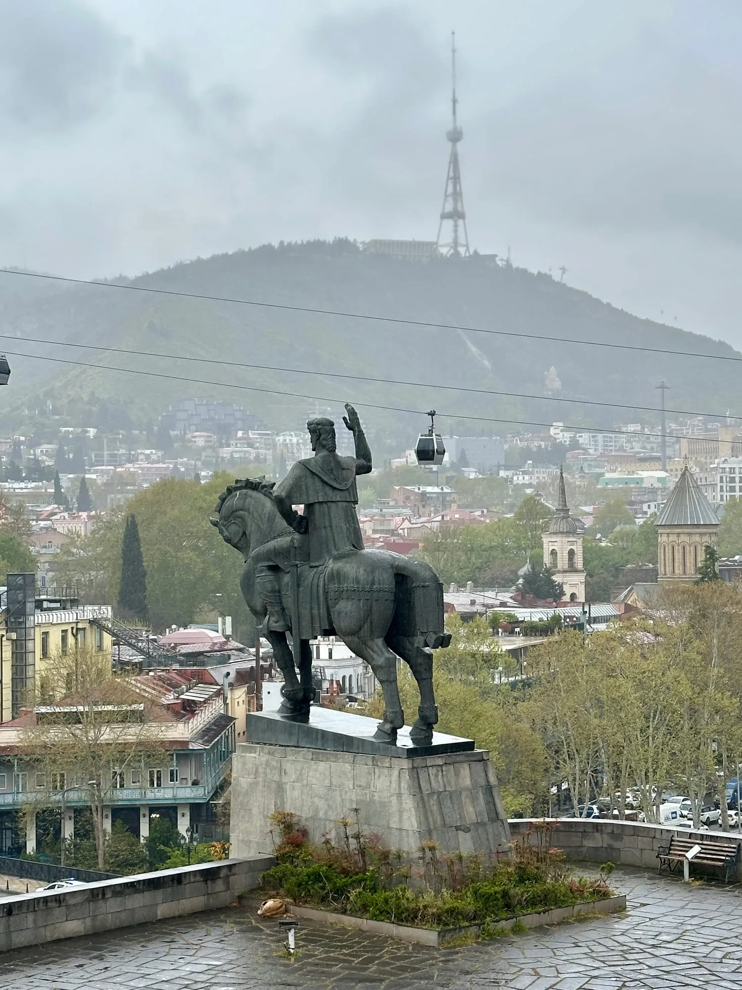 Reiterstandbild mit erhobenem Arm vor der hügeligen Stadt Tbilisi, dahinter eine Seilbahnkabine.