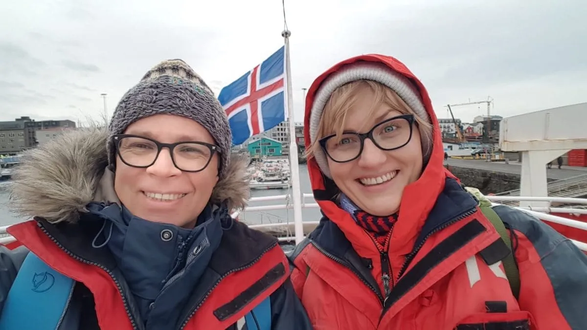 Iwona und Lukas in Winterjacken auf einem Schiff, dahinter eine isländische Flagge