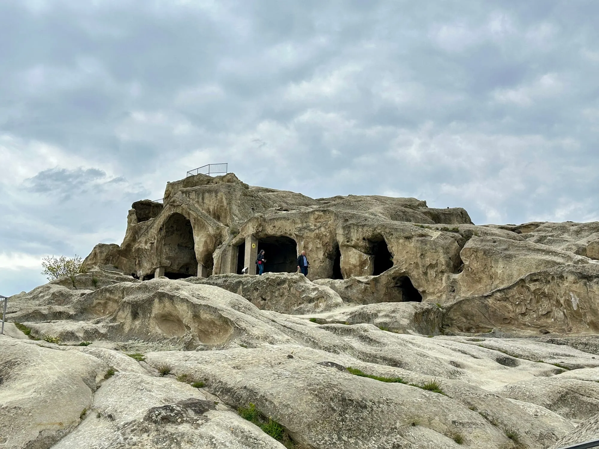 In den Fels gehauene Höhlenanlage von Uplisziche mit Besuchern und einem Metallgeländer unter wolkenverhangenem Himmel.